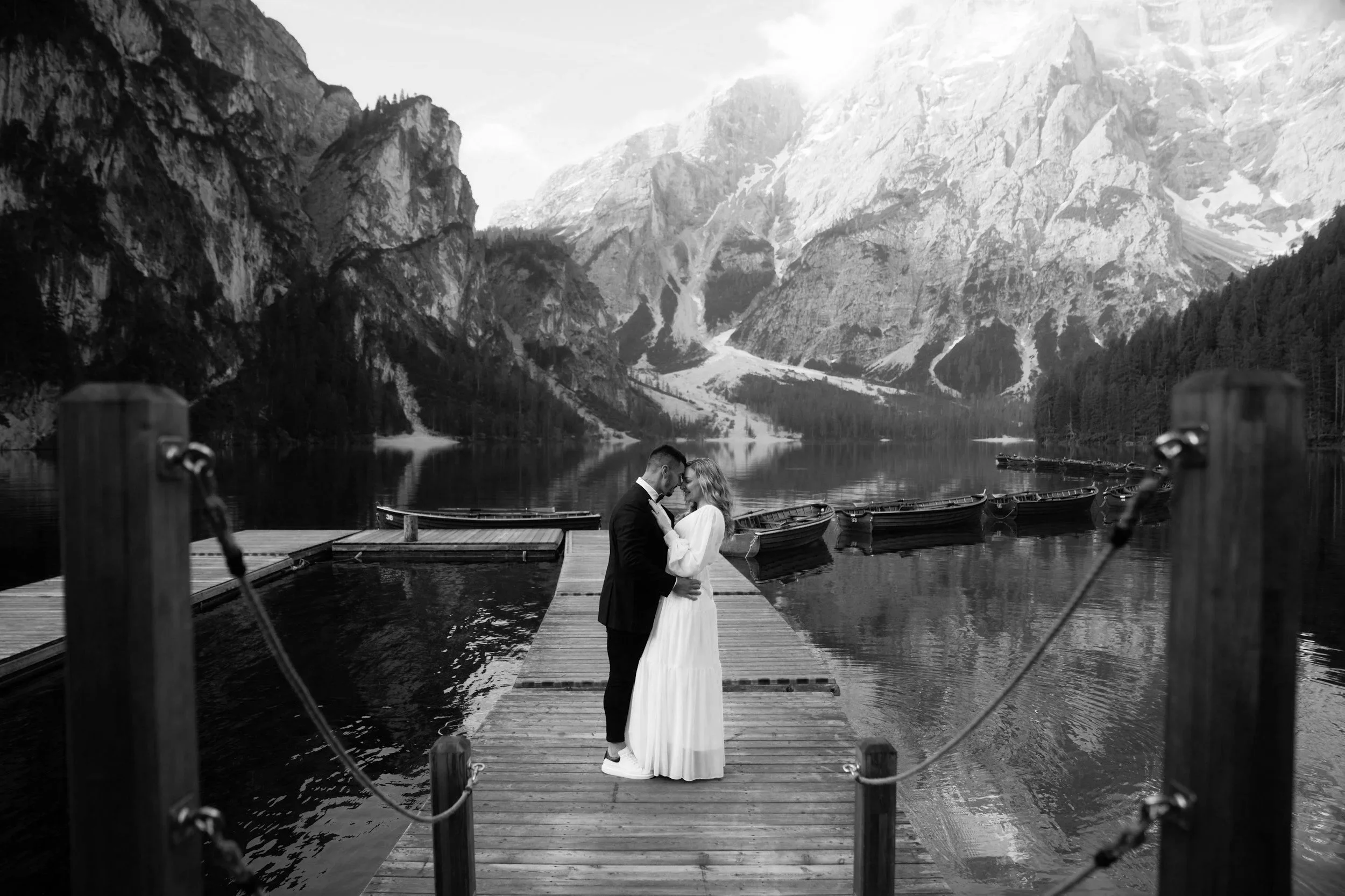 Ein berührendes Brautpaar steht auf einem Steg am See in den Bergen, exchanging liebevolle Blicke, umgeben von einer malerischen Landschaft mit hohen Bergen im Hintergrund, in Schwarz-Weiß.
