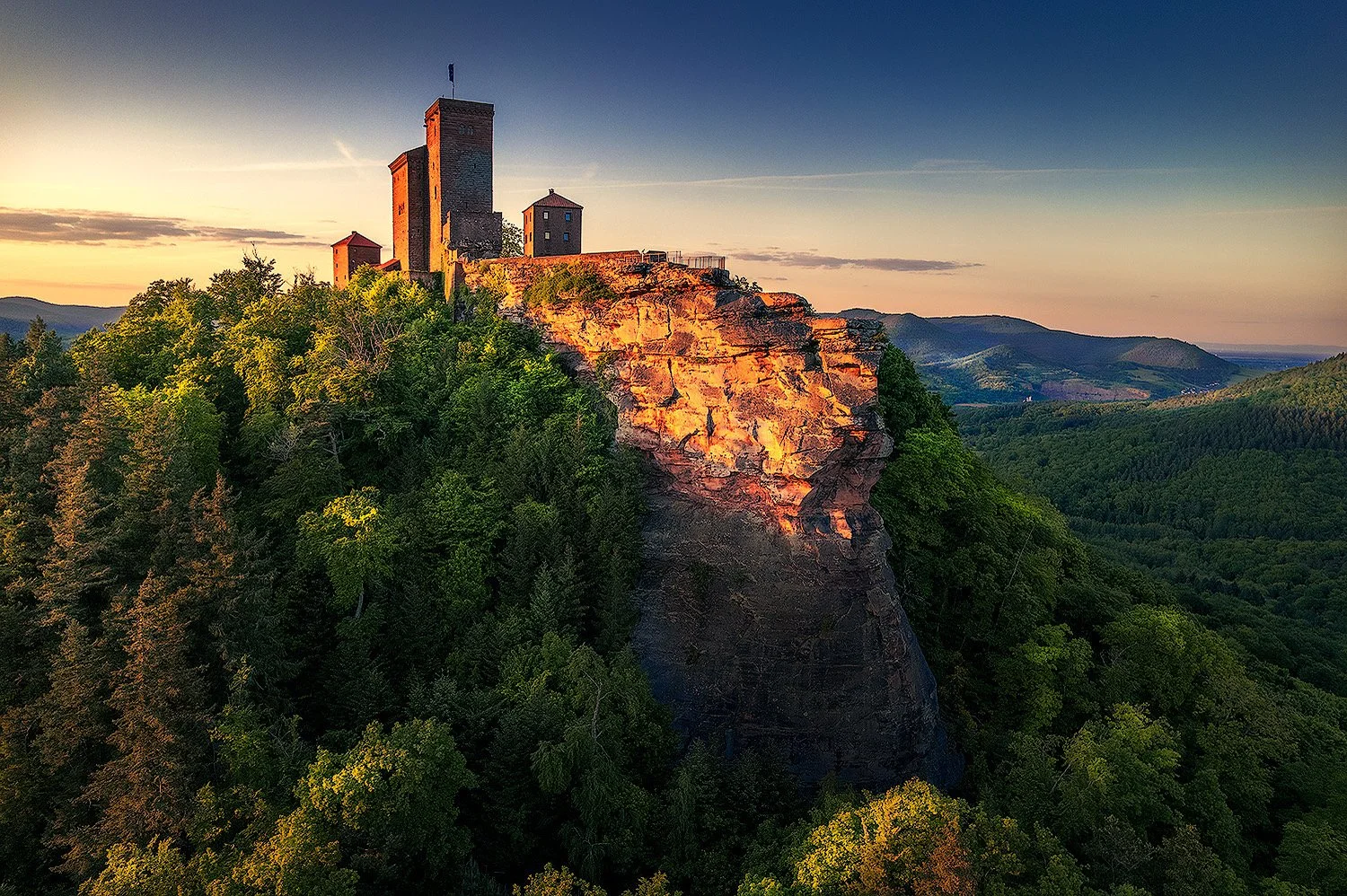 Burg auf einem Felsen am Abend, umgeben von einem Wald, mit Hügeln im Hintergrund.