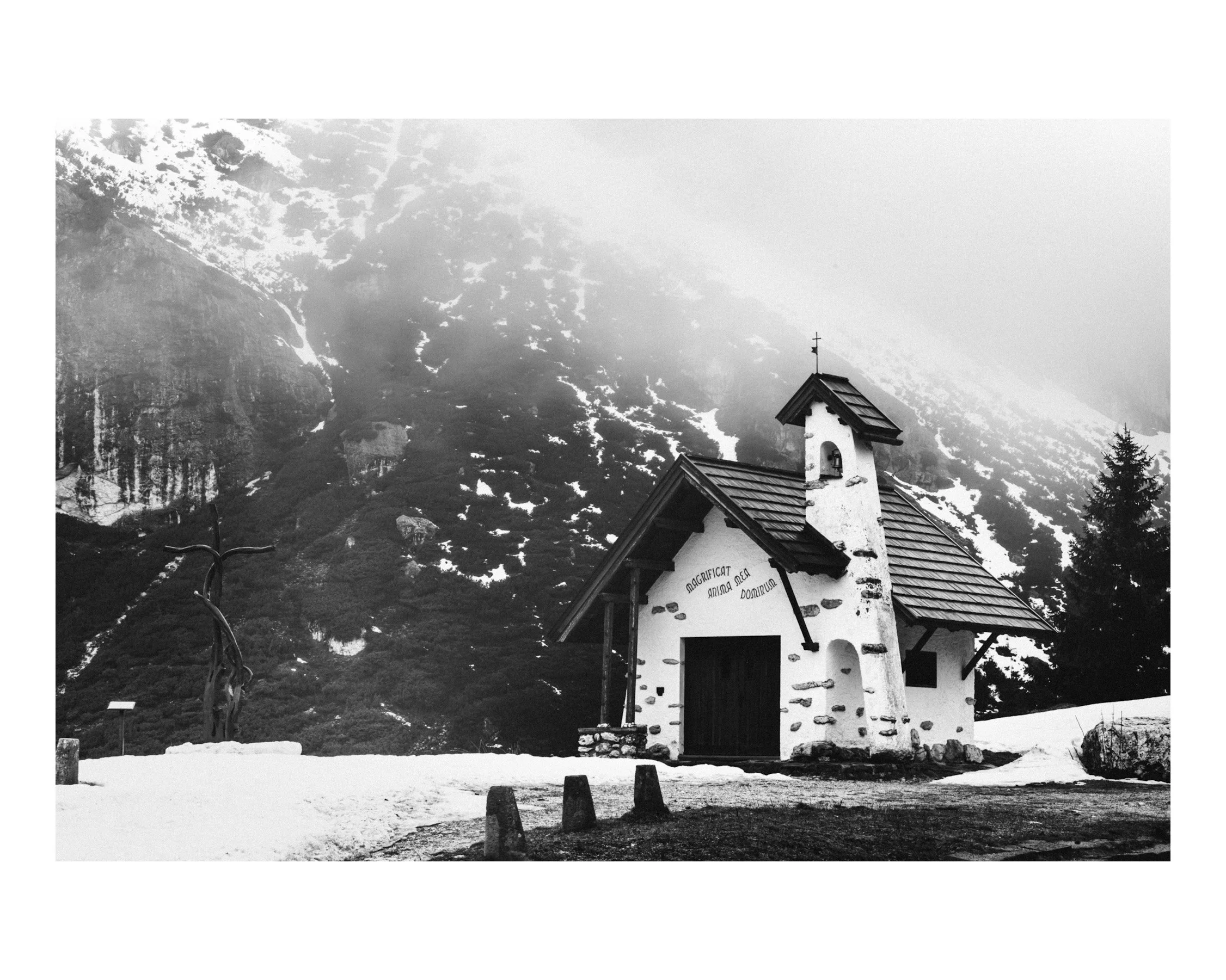 Kleine weiße Kapelle in verschneiter Berglandschaft mit bewaldeten Bergen im Hintergrund, neblig und winterlich.