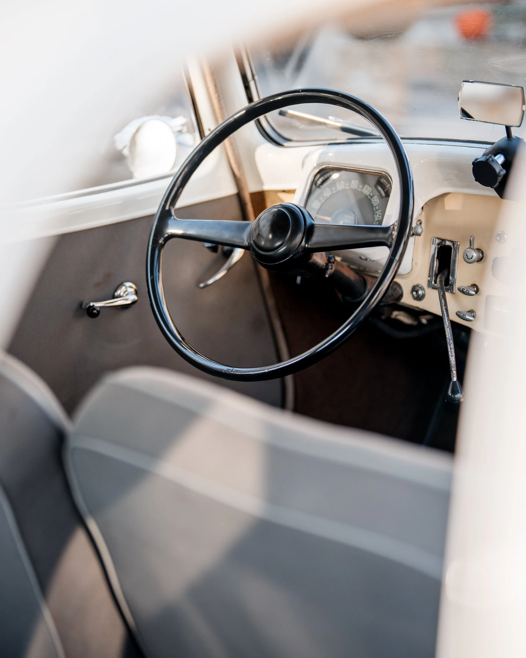 Innenraum eines Vintage-Autos mit schwarzem Lenkrad, Tachometer, Schaltknüppel, und Armaturenbrett in cremefarbenem Design.