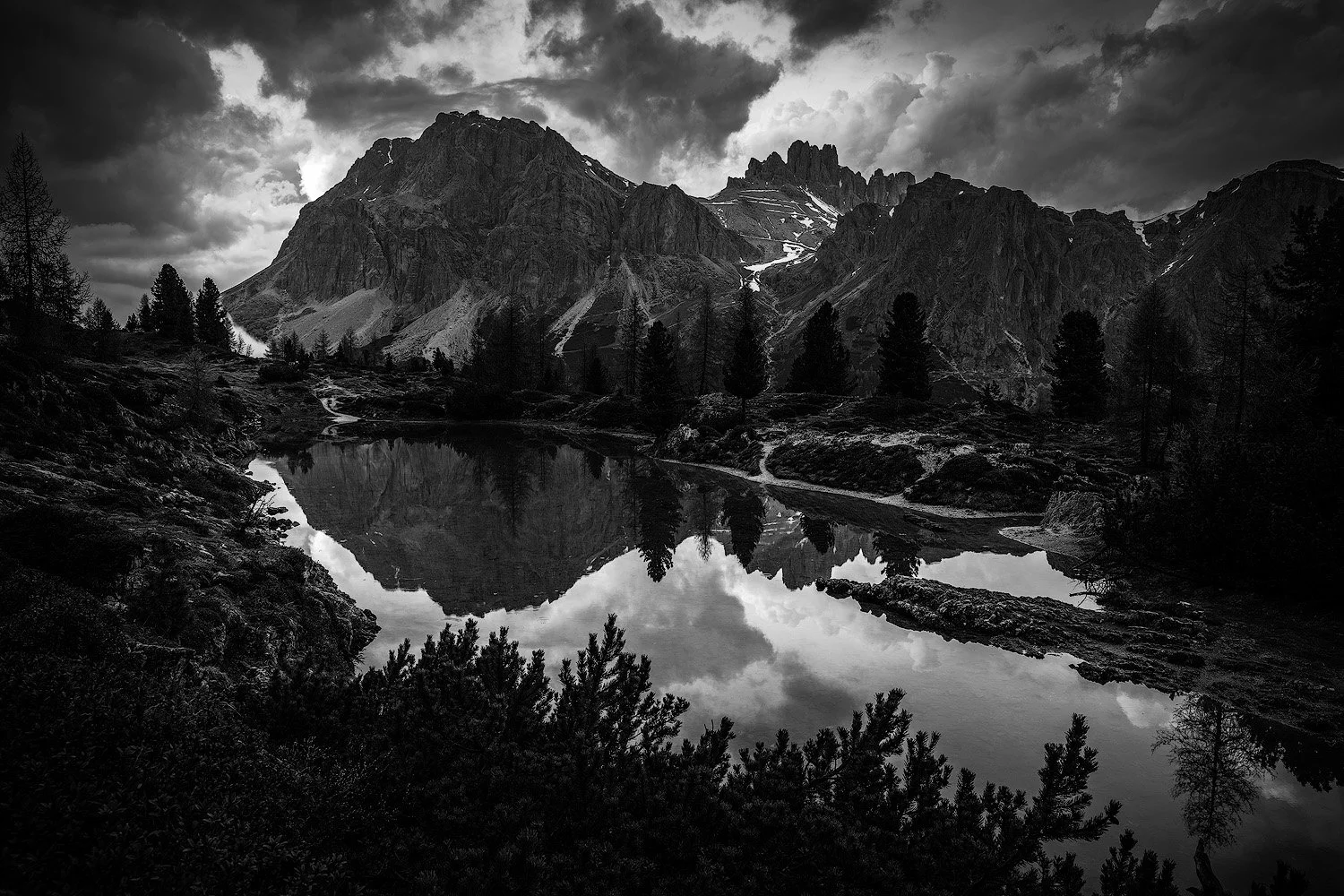 Schwarze und weiße Aufnahme eines Bergsees mit reflektierenden Bergen und dunklen Wolken am Himmel, umgeben von Bäumen und Pfaden.