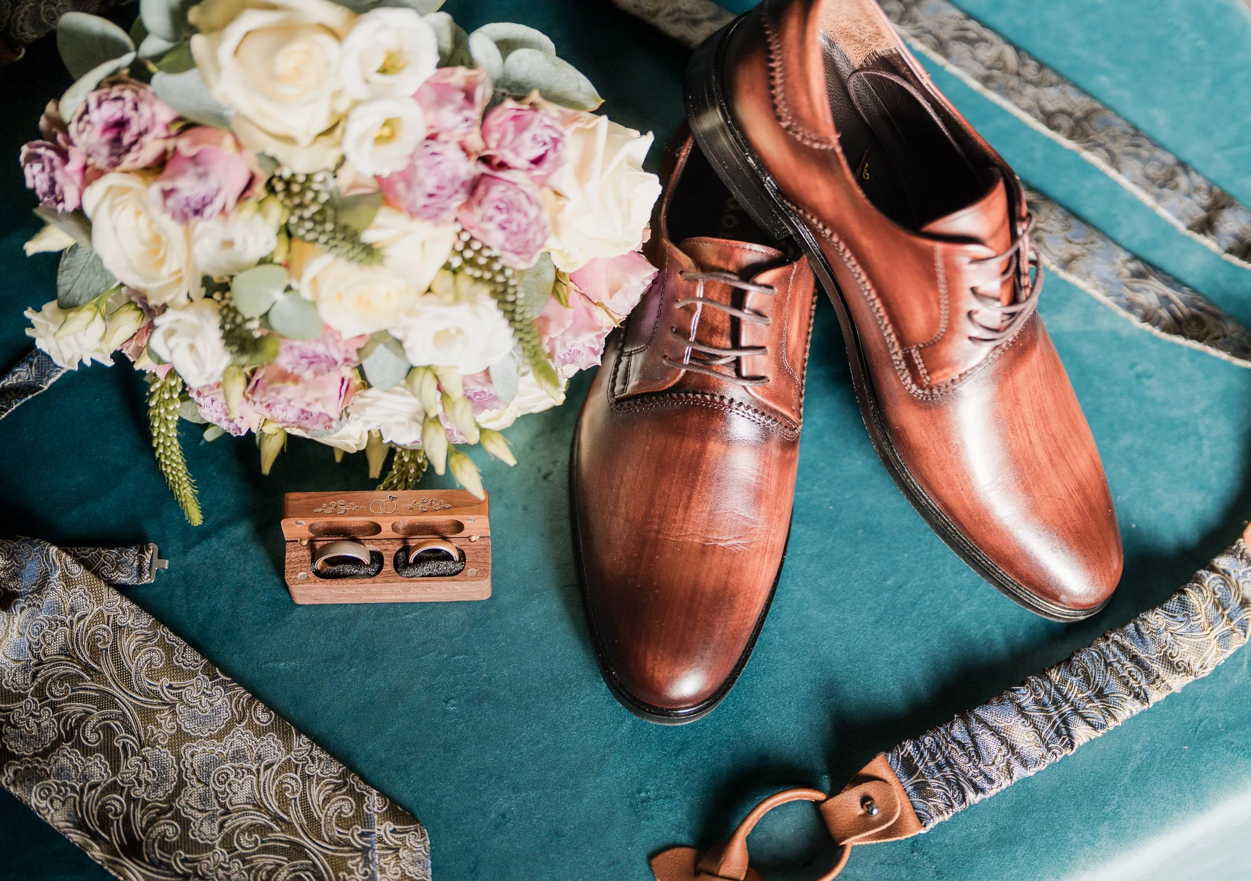 Braune Lederschuhe, Blumenstrauß mit weißen, rosa und lila Blumen, Eheringe in einer Holzschatulle, Stoffgürtel mit Muster auf grünem Stoff