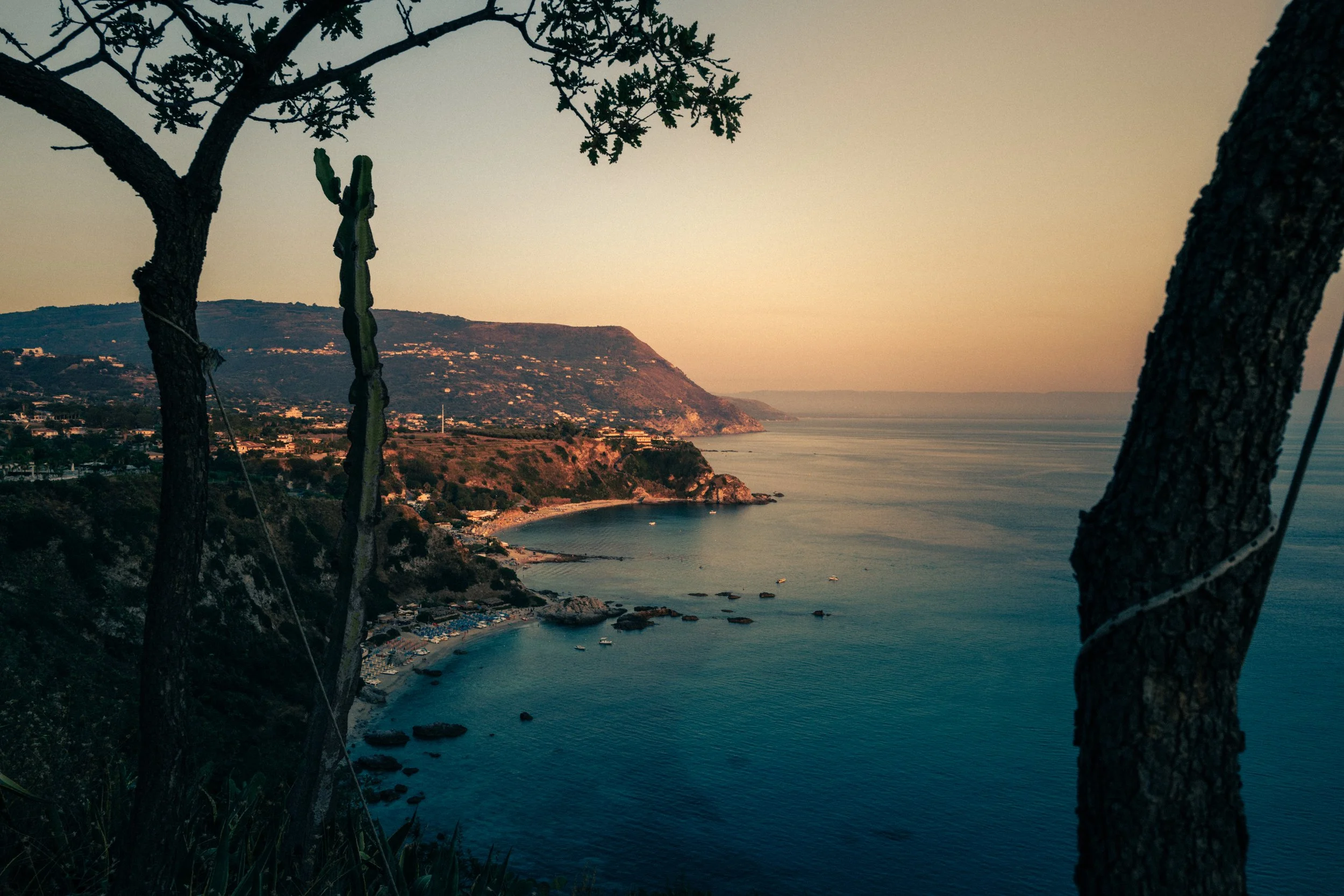 Blick auf eine Küstenlinie mit Bäumen im Vordergrund, Wasser, Strand und Hügel im Sonnenuntergang. Calabria, Italia, Capo Vaticano.