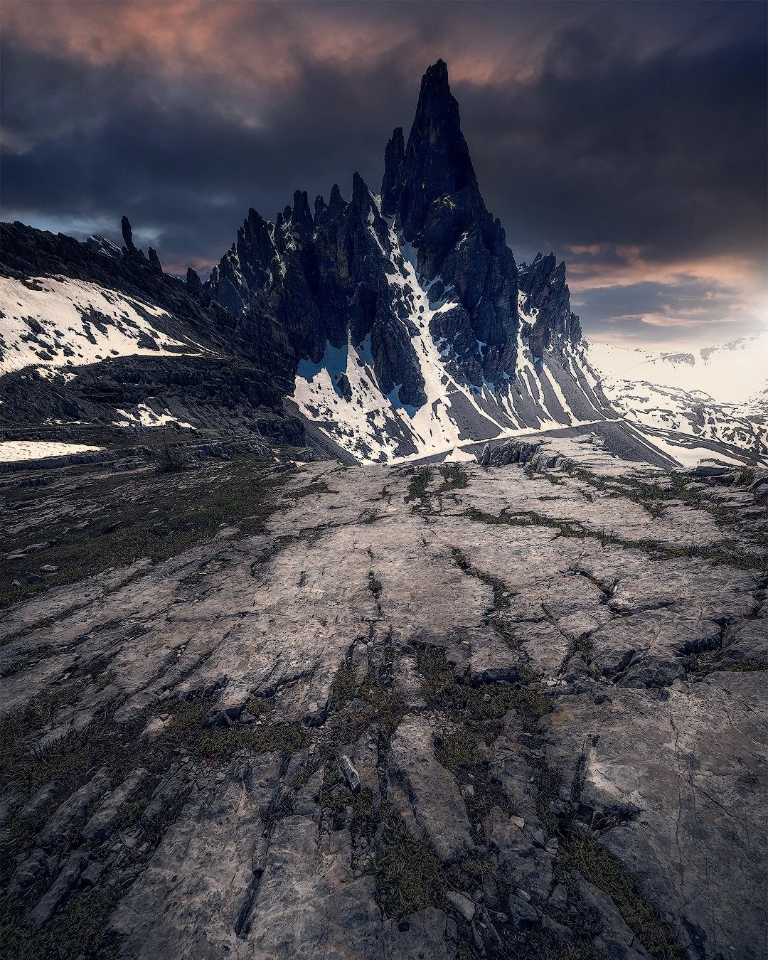 Berglandschaft mit steiler, spitz zulaufender Bergspitze, Schneefeldern und einer felsigen, unebenen Oberfläche im Vordergrund bei Sonnenuntergang