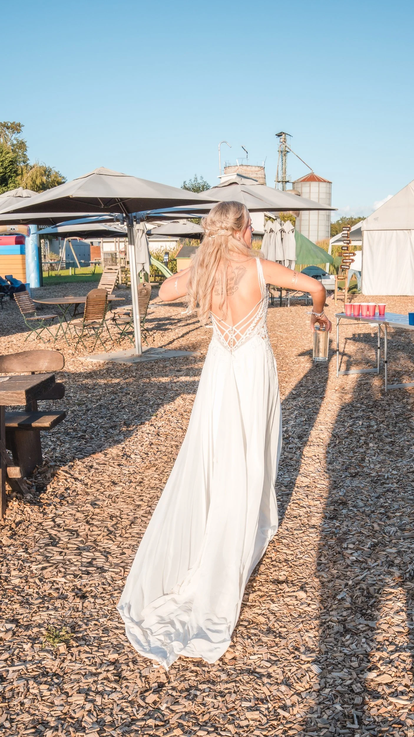 Frau in weißem Brautkleid mit offenem Rücken, mit langen, blonden Haaren, auf einem Freigelände mit Sonnenschirmen, Holzliegen, Zelten und einem Spielplatz im Hintergrund, bei Sonnenuntergang.