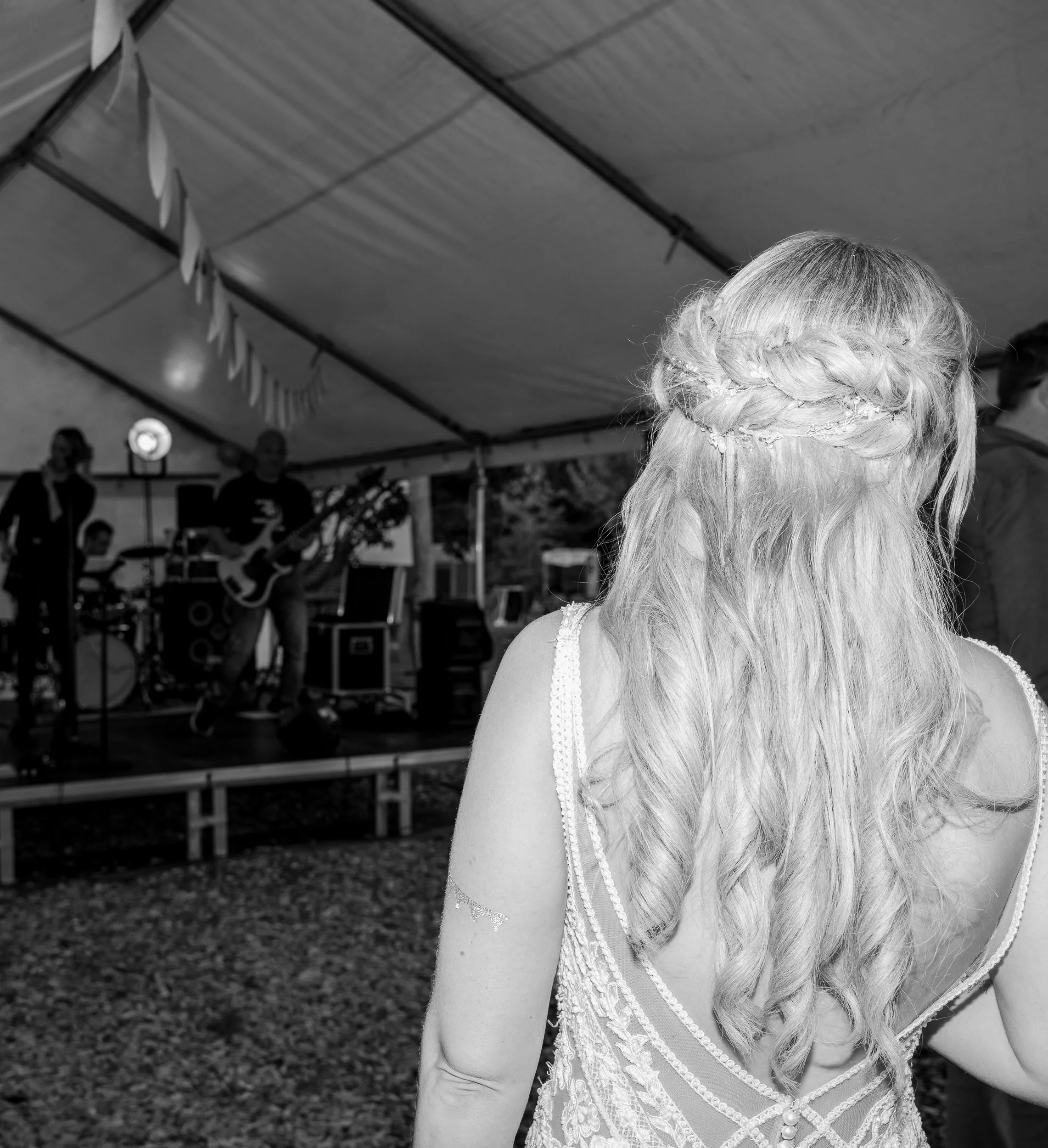 Frau mit langen, gelockten Haaren und dezentem Brautkleid, blickt auf eine Band auf der Bühne im Zelt bei einer Feier oder Hochzeit.