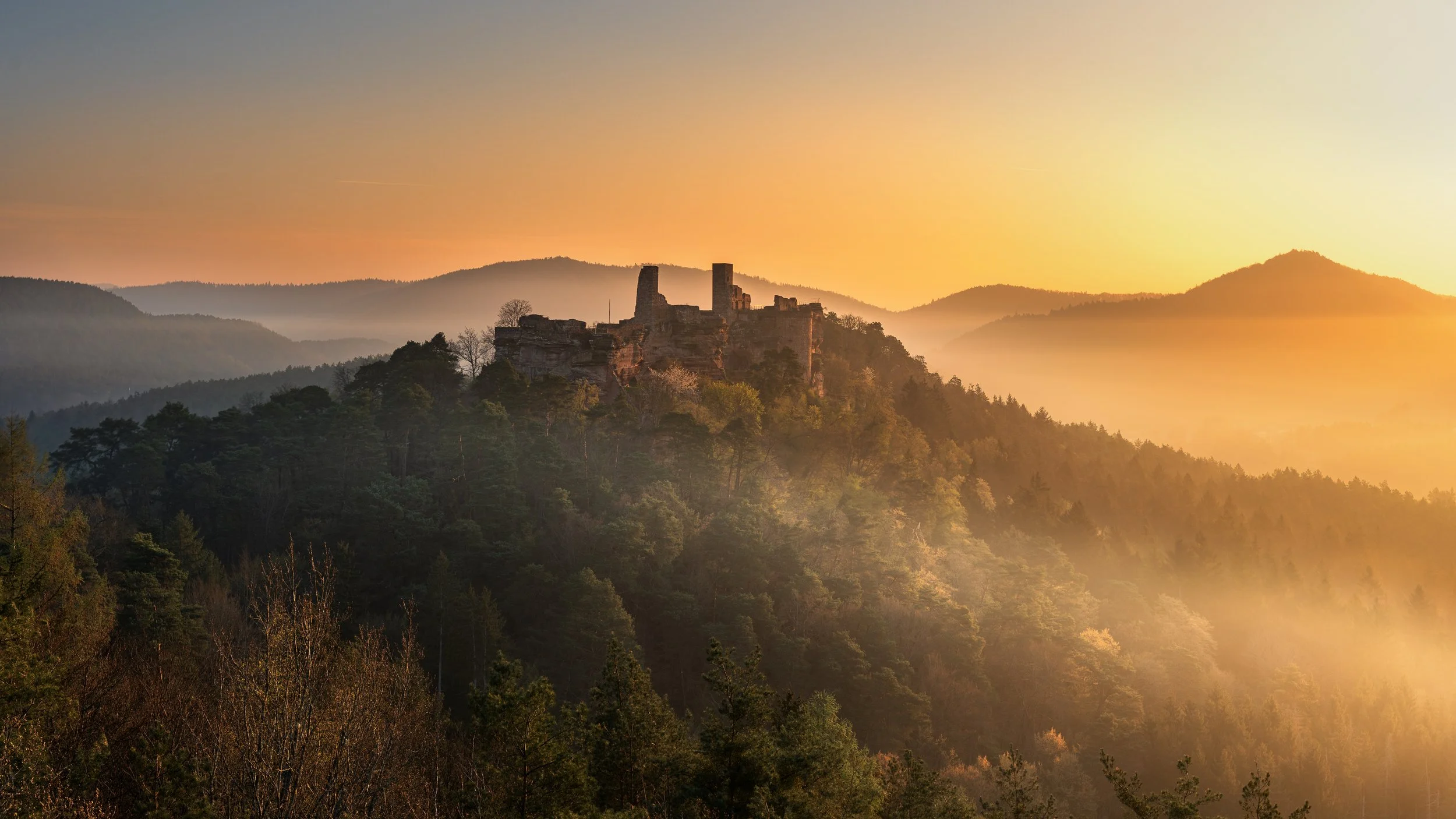 Burg auf einem Hügel während eines Sonnenuntergangs, umgeben von bewaldeten Hügeln und Nebel. Dahner Felsenland.