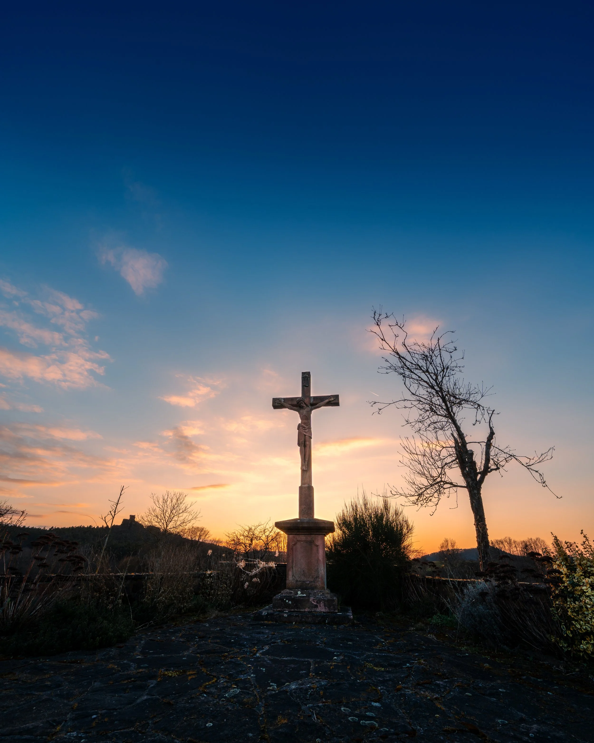 Christus-Kreuz auf einem Steinpodest bei Sonnenuntergang, umgeben von Büschen und Bäumen, mit einem bewölkten Himmel im Hintergrund.