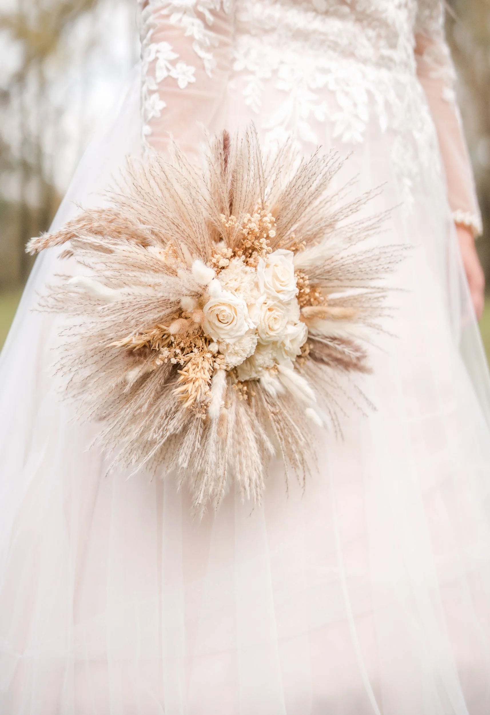 Braut trägt ein Brautkleid mit Spitzenmuster, hält einen Blumenstrauß mit weißen Rosen und getrockneten Gräsern.