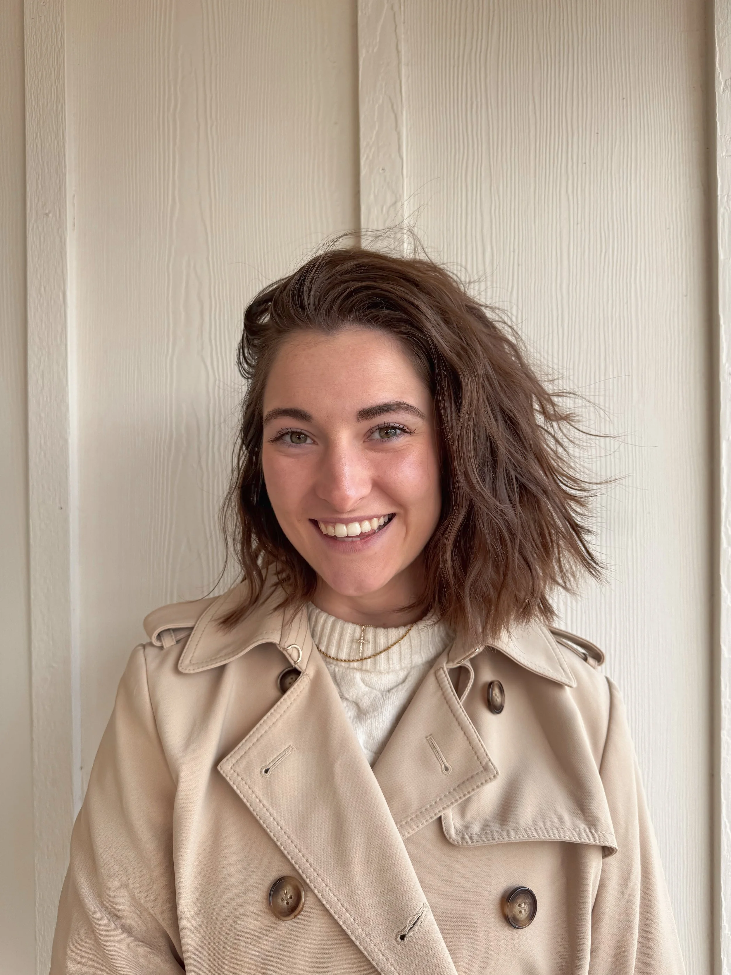 A woman with shoulder-length wavy brown hair smiling, wearing a beige trench coat and a white sweater, standing in front of a light-colored wooden wall.