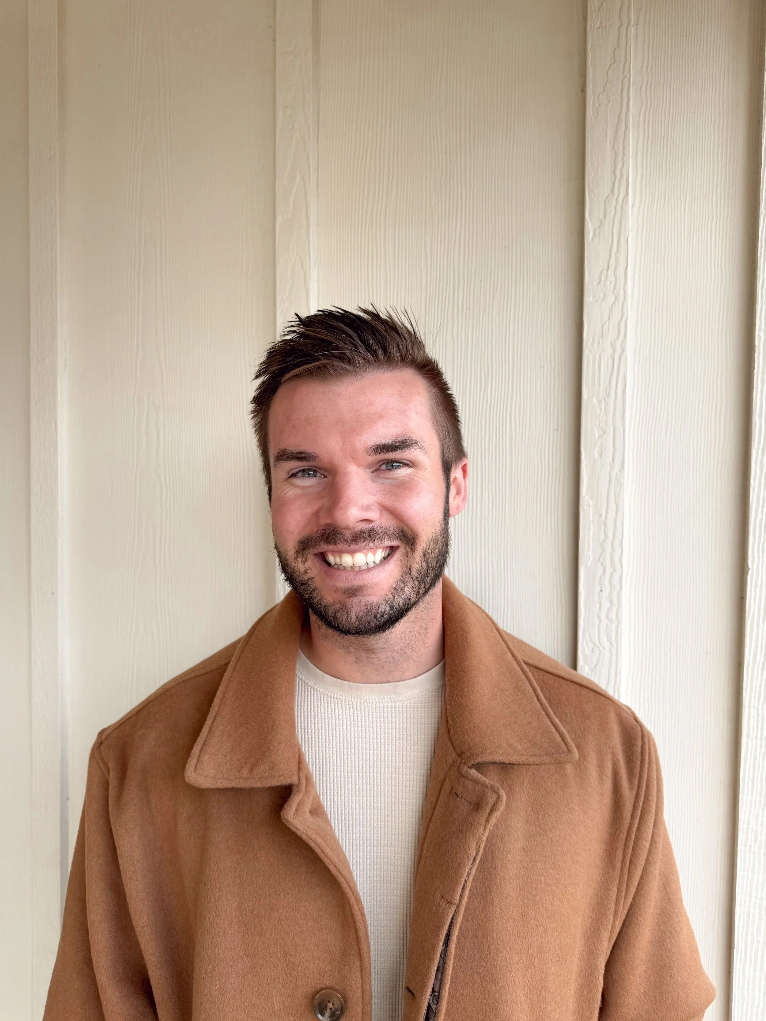 A smiling young man with dark hair and a beard, wearing a tan coat and a white shirt, standing against a wooden wall.