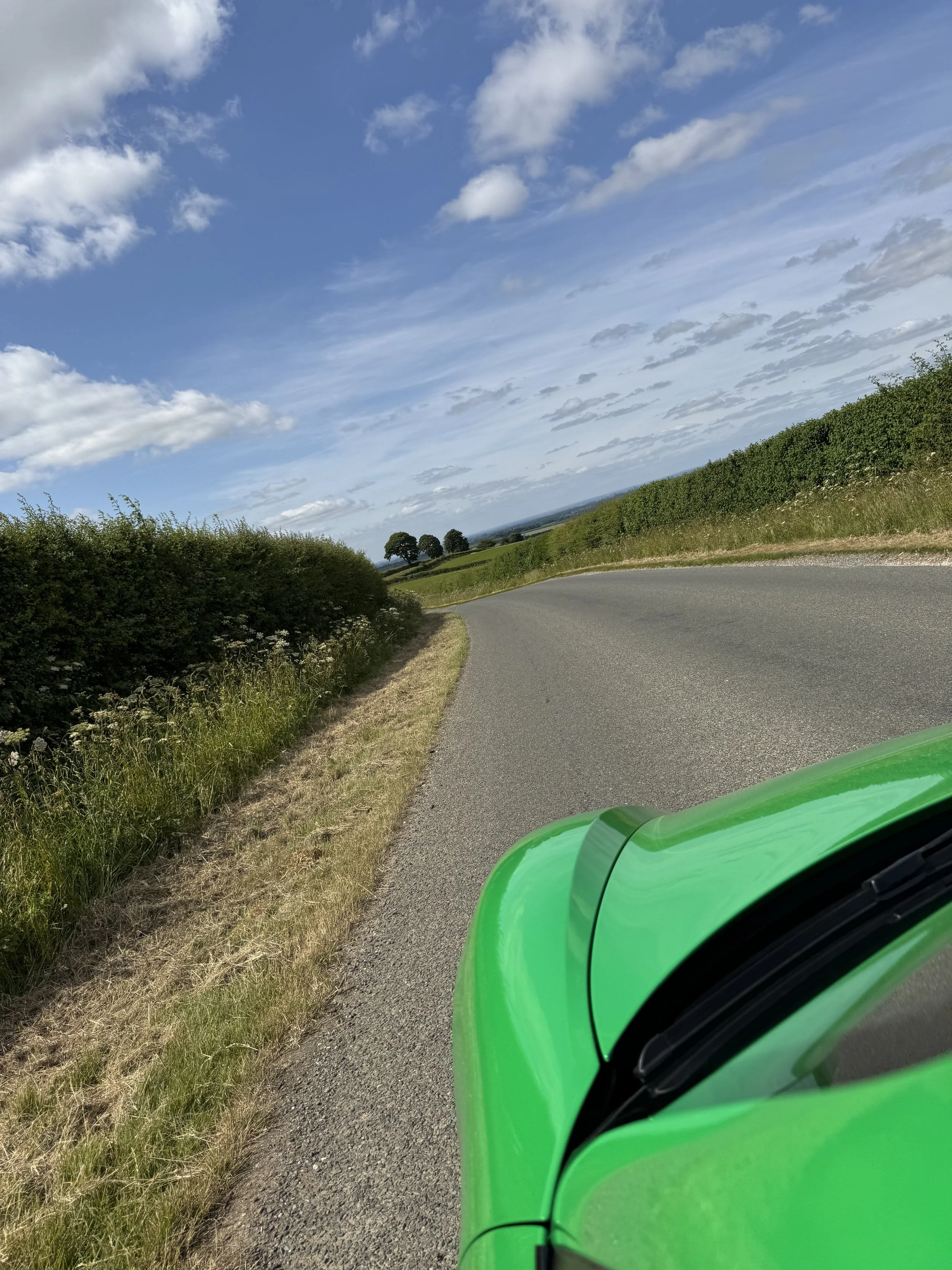 Part of a bright green sports car on a winding country road with green hedges on either side and a partly cloudy blue sky overhead.