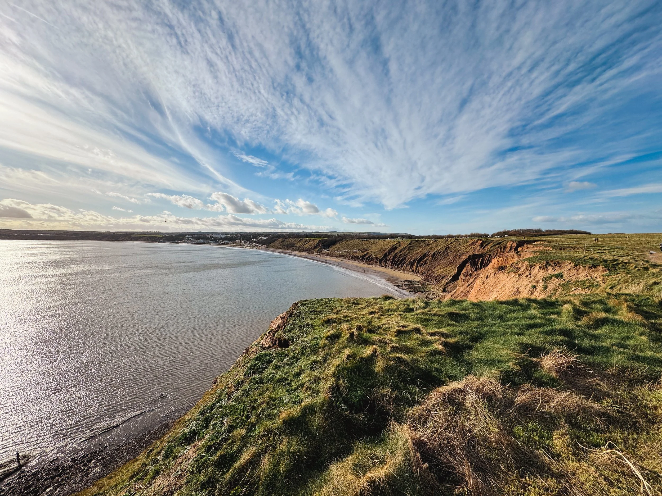 Cliffside view of coastline with grassy area in foreground, sandy beach, calm water, and a sky with scattered clouds.