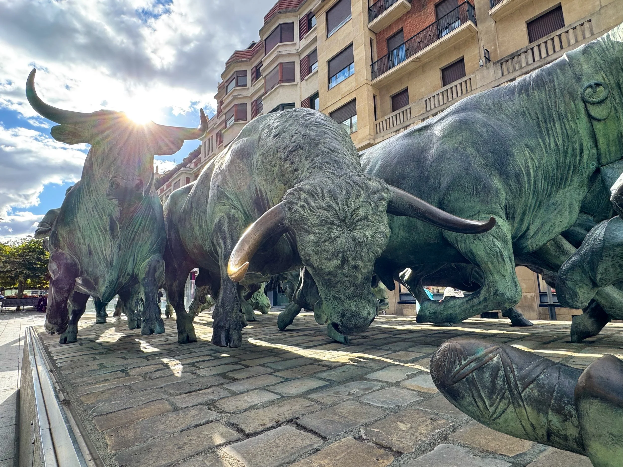 Bronze sculptures of a herd of bison on a cobblestone platform in an urban setting, with a tall building in the background and the sun shining through the clouds.