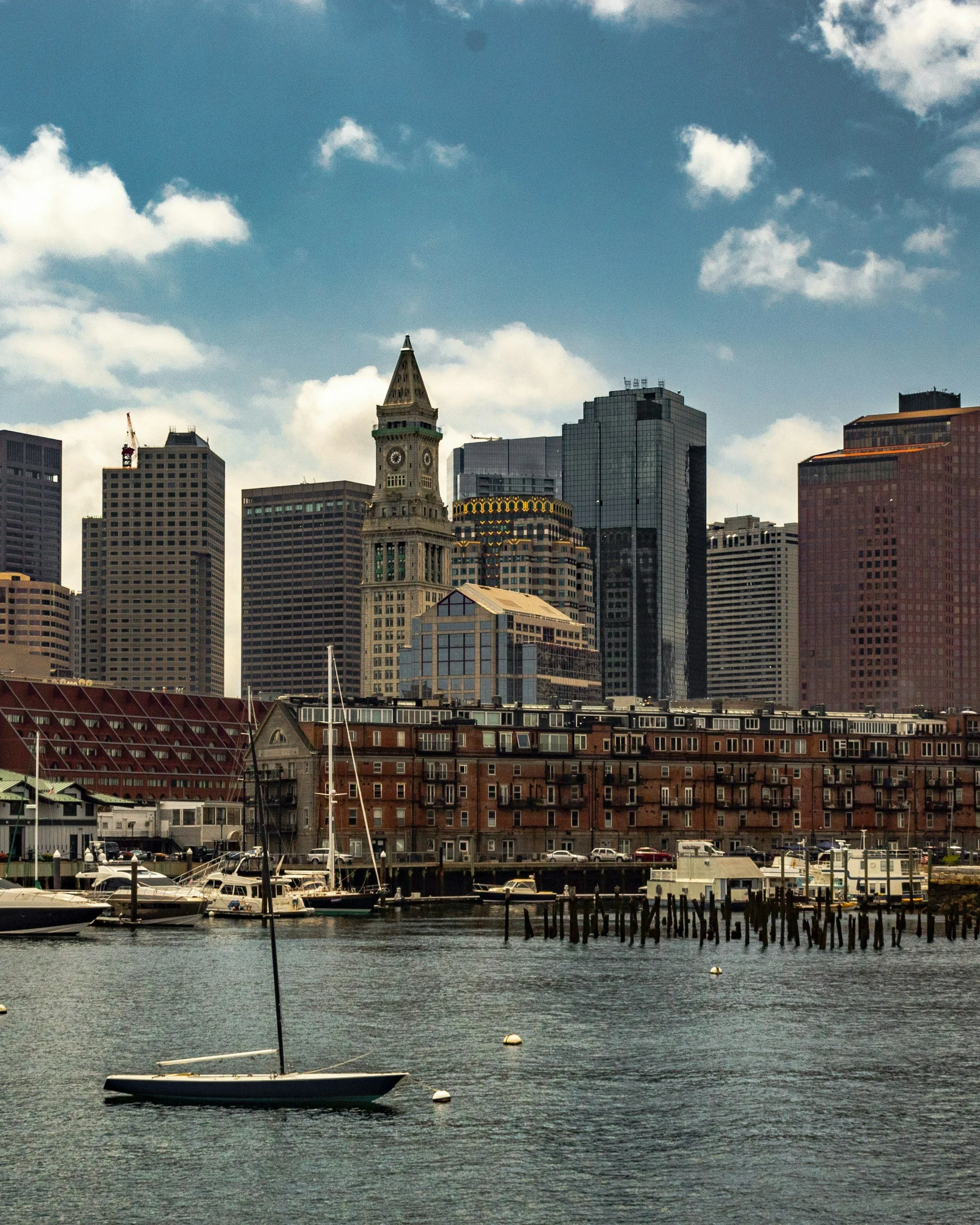A city skyline with tall skyscrapers, a historic clock tower, boats docked in the water, and a partly cloudy sky.