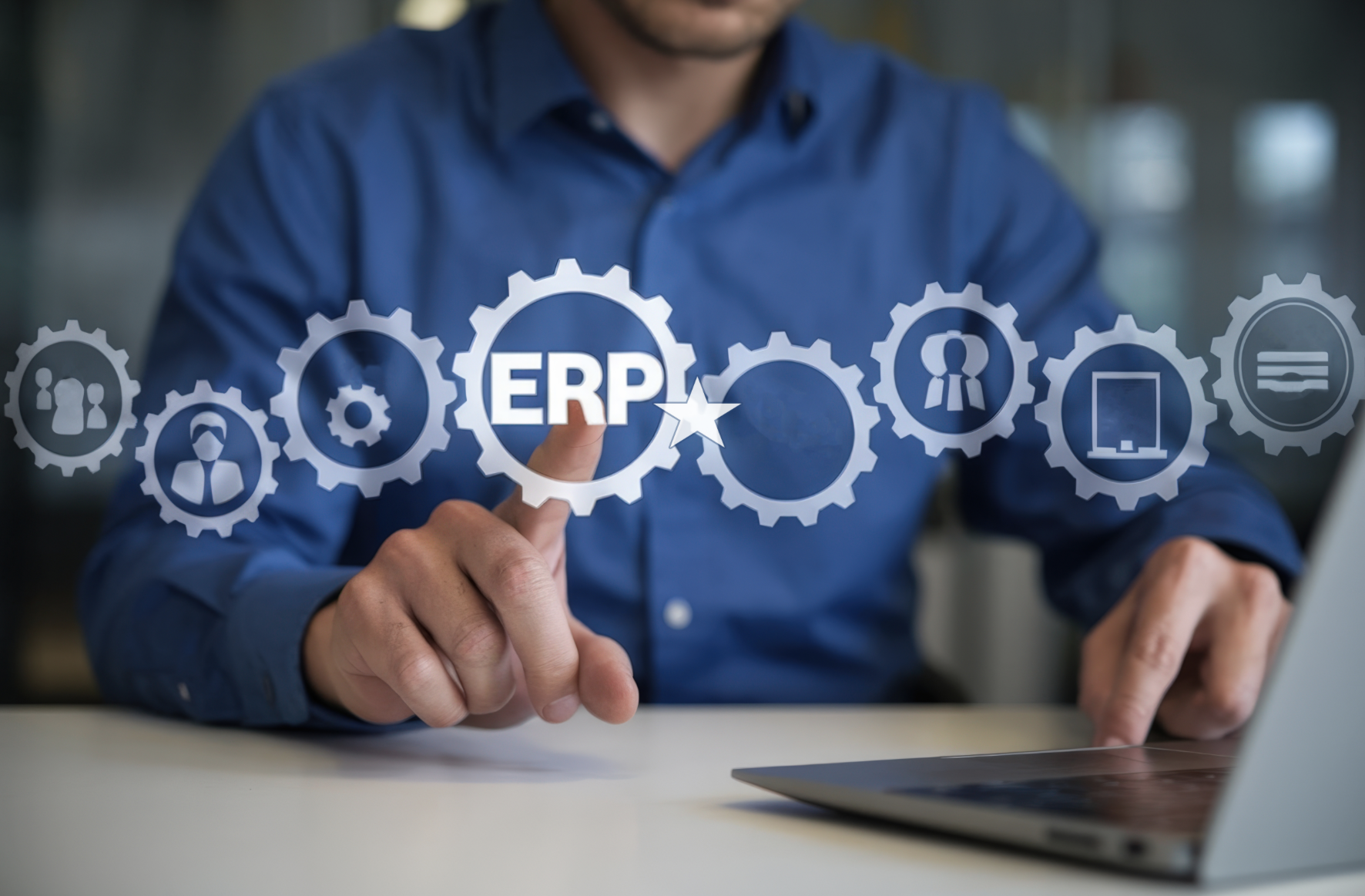 A person in a blue shirt interacts with digital icons representing ERP systems on a transparent screen in front of them, with a laptop on the table.