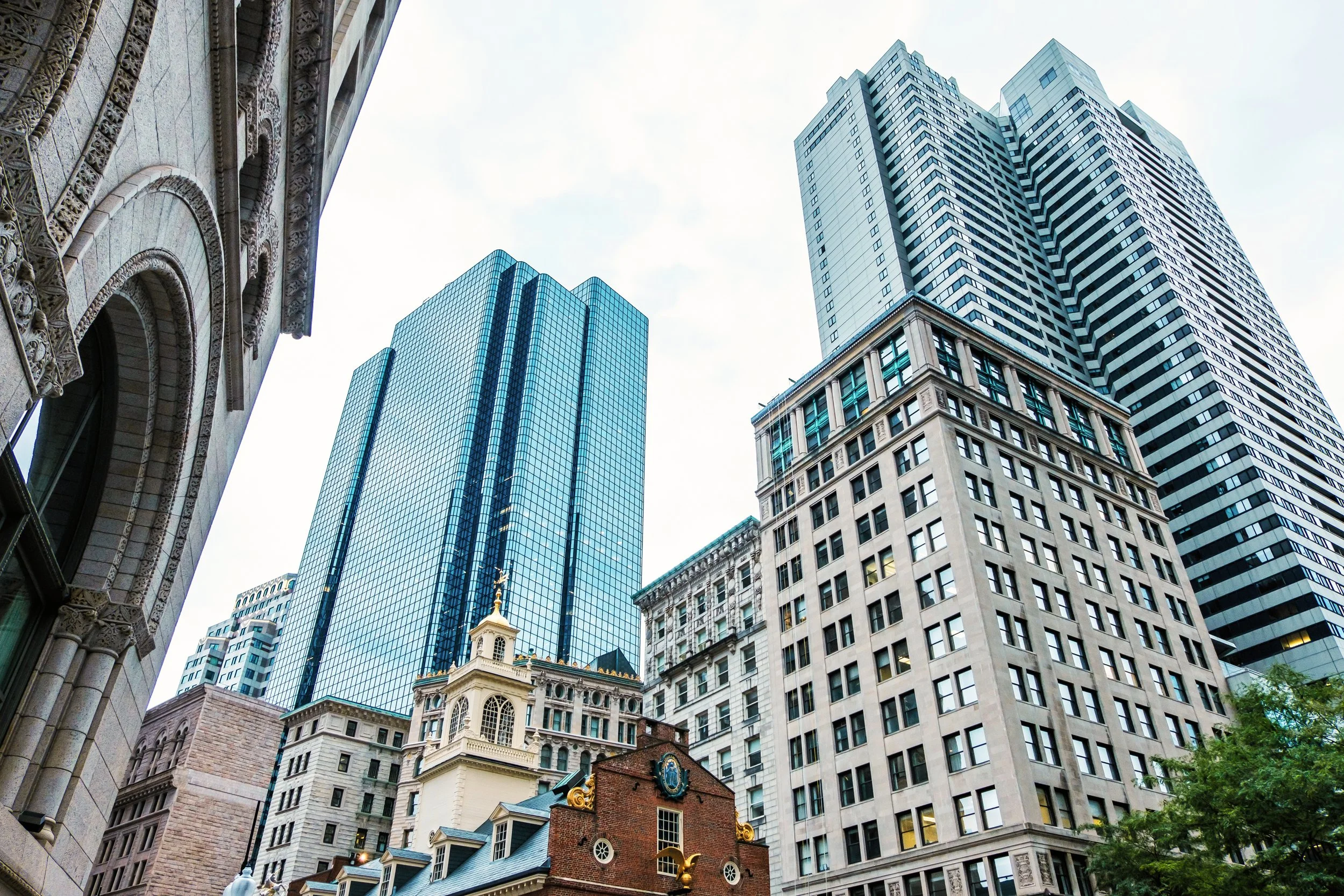 Low-angle view of tall modern skyscrapers and historic buildings in a city, with a partly cloudy sky in the background.