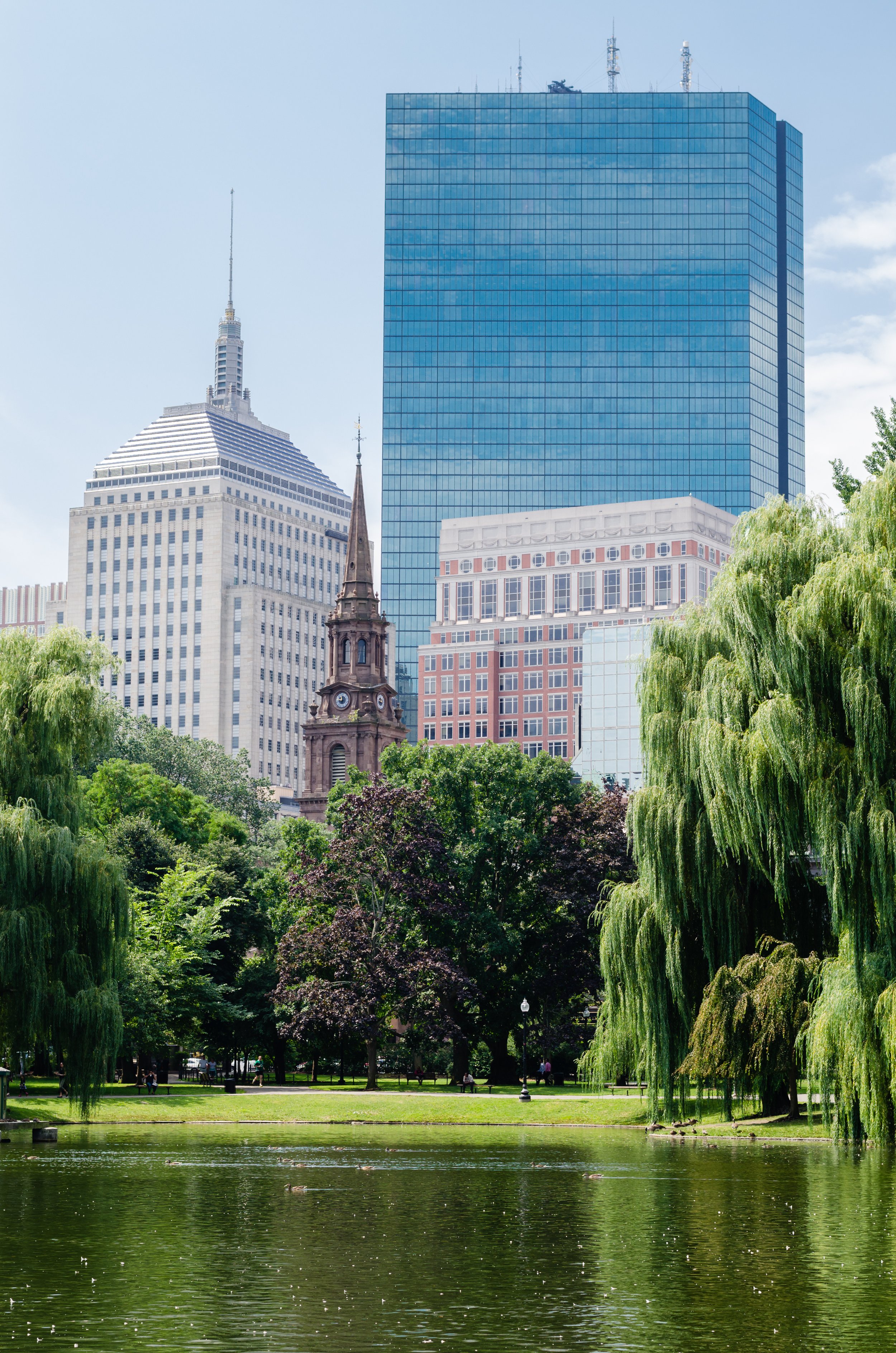 A city park with green trees and a pond in the foreground, and tall skyscrapers including a modern glass building, a historic church with a clock tower, and a variety of other buildings in the background.