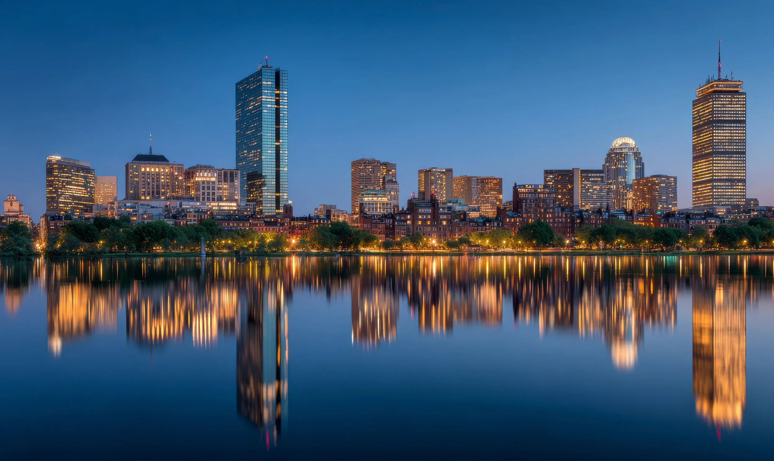 City skyline of Boston at dusk with illuminated buildings reflecting on calm water in the foreground.