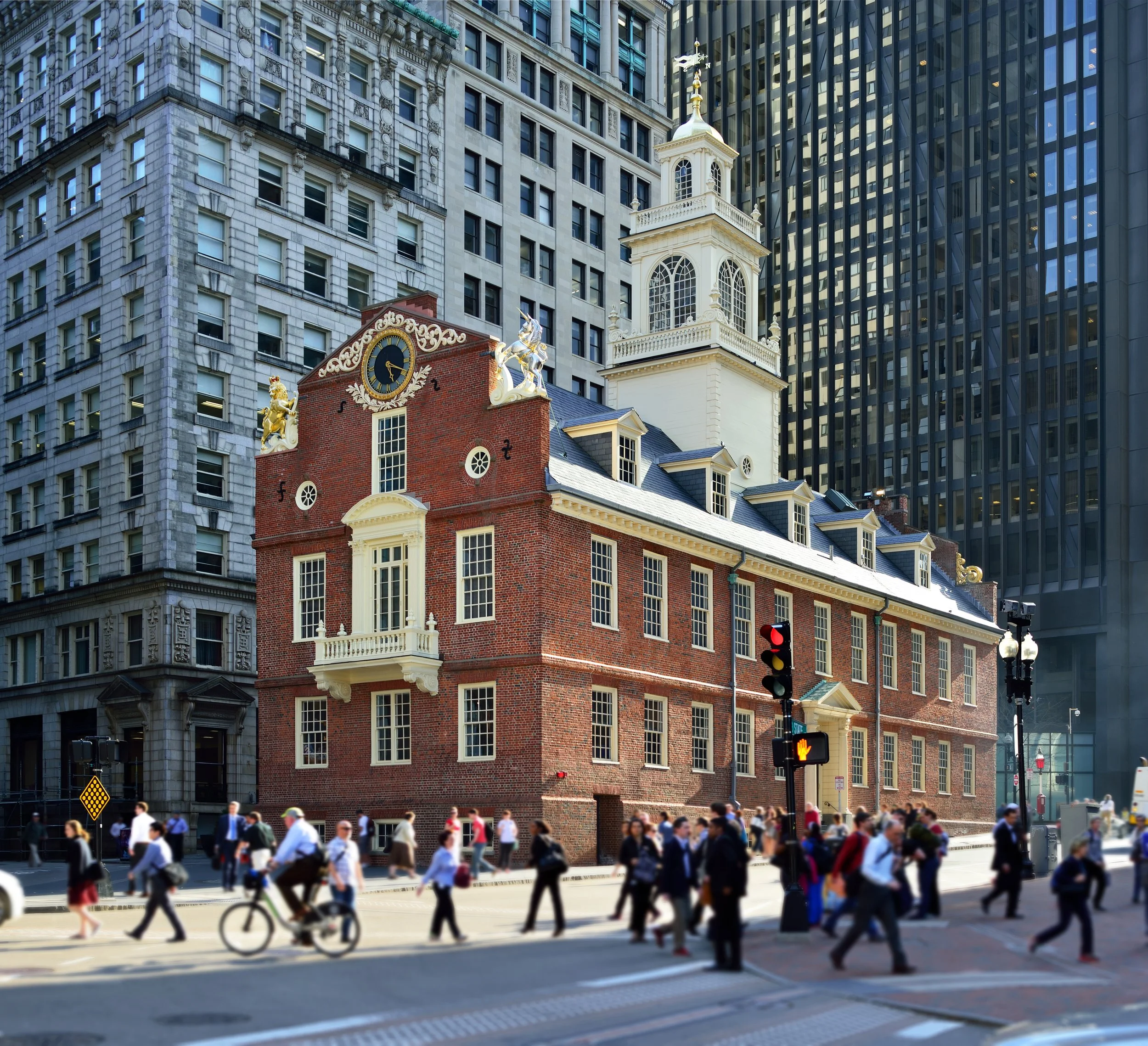 The image shows an historic building with brick and white decorative accents, set amidst modern skyscrapers. The building features a clock and decorative sculptures on the roof. Pedestrians and cyclists are crossing the street in front of the building, and traffic lights are visible.