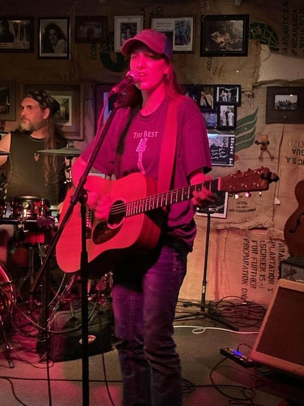 A woman playing an acoustic guitar and singing into a microphone at a live music venue, with a drummer in the background.