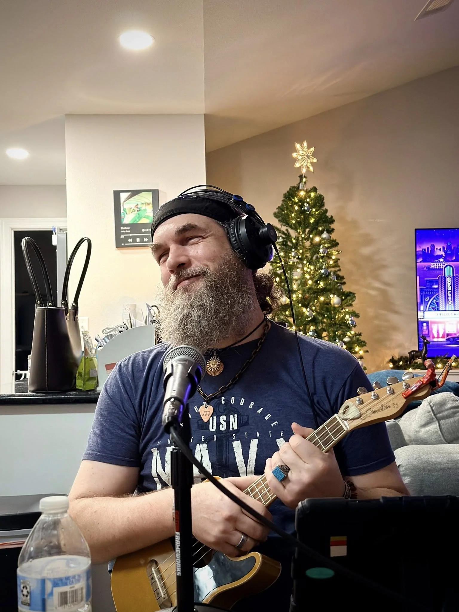 A bearded man wearing headphones and a dark cap playing a ukulele near a microphone indoors, with a decorated Christmas tree and a TV screen in the background.