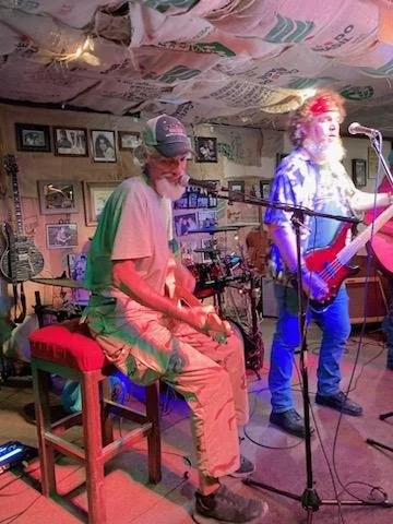Two musicians performing on a small stage inside a cozy venue with framed pictures on the wall. One is sitting on a red stool, wearing a hat and casual clothes, playing a percussion instrument. The other is standing, wearing a bandana and sunglasses, singing into a microphone while playing an acoustic guitar. The ceiling is decorated with hanging newspaper.