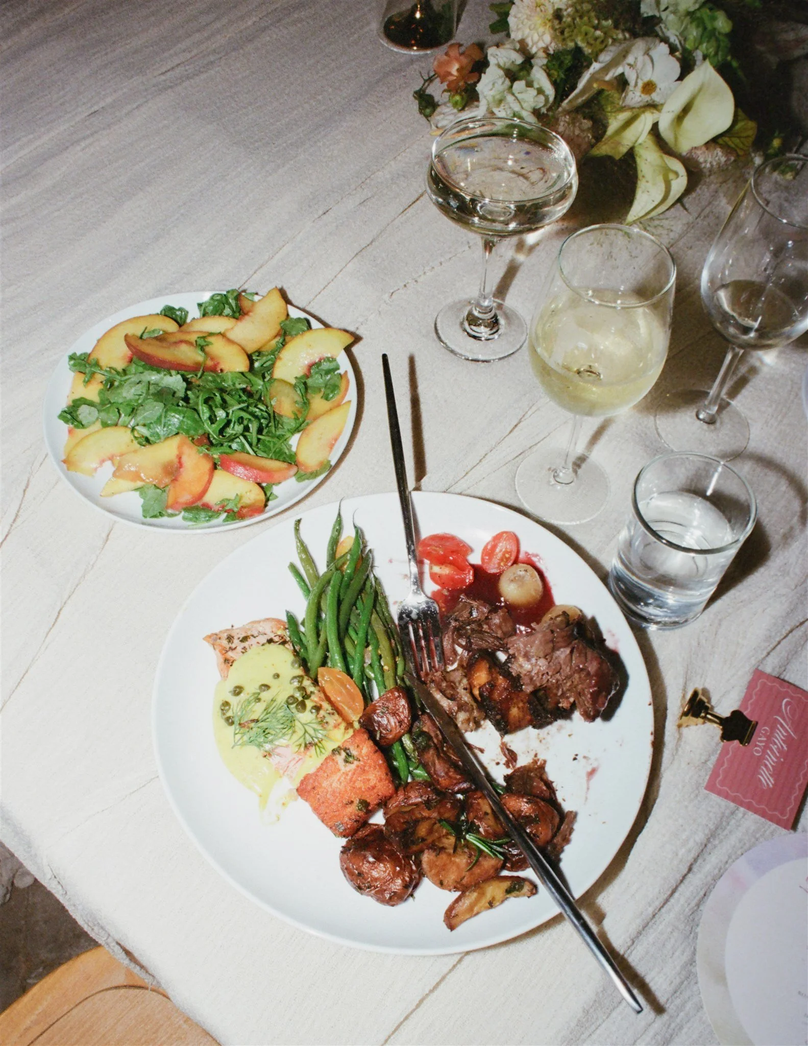 A dinner table with a white plate of salad with peach slices and greens, a plate of cooked meats with green beans and roasted vegetables, and glasses of water, white wine, and champagne. There are also some flowers in a vase in the background.