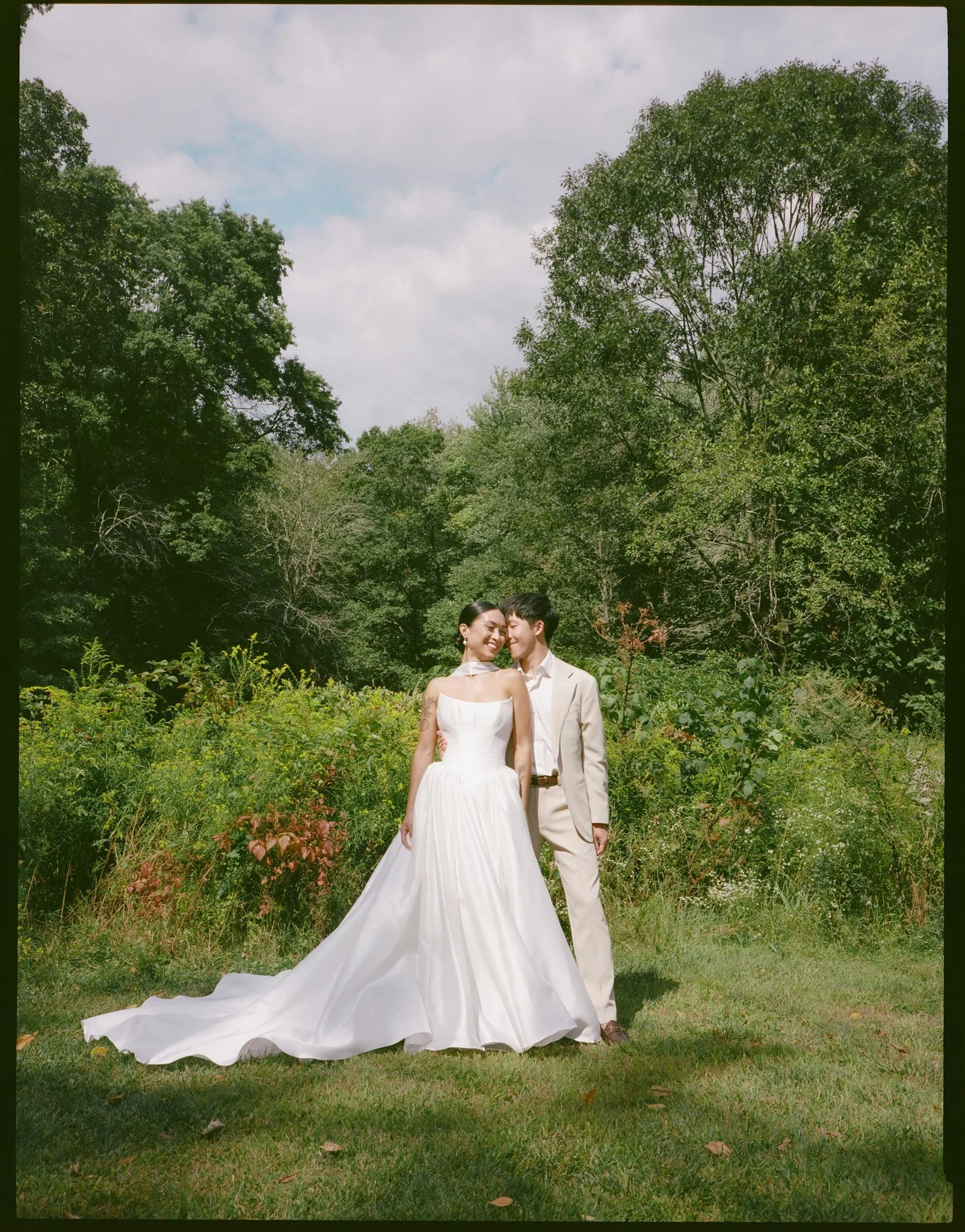 A bride and groom smiling and standing outdoors in a grassy area with trees in the background, the bride wearing a white gown and the groom in a light beige suit, during a wedding photoshoot.