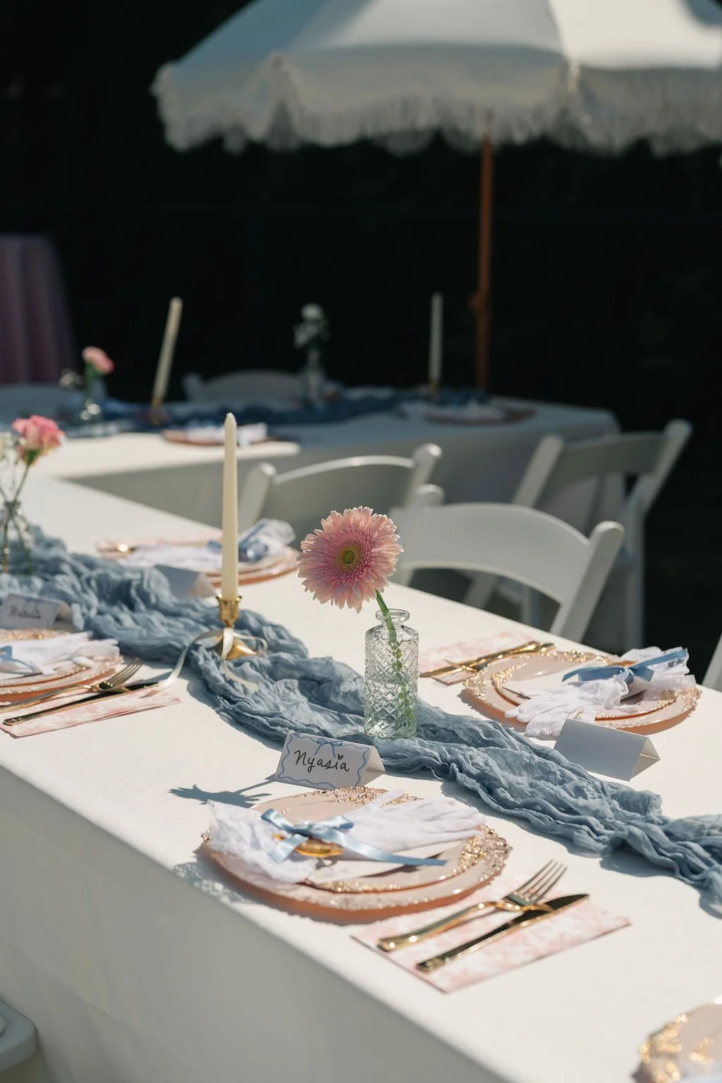 A decorated outdoor dining table with a white tablecloth, featuring a pink flower in a glass vase, gold cutlery, and white napkins tied with light blue ribbons. There are also white candles in gold holders, place cards, and a gray fabric runner. A be