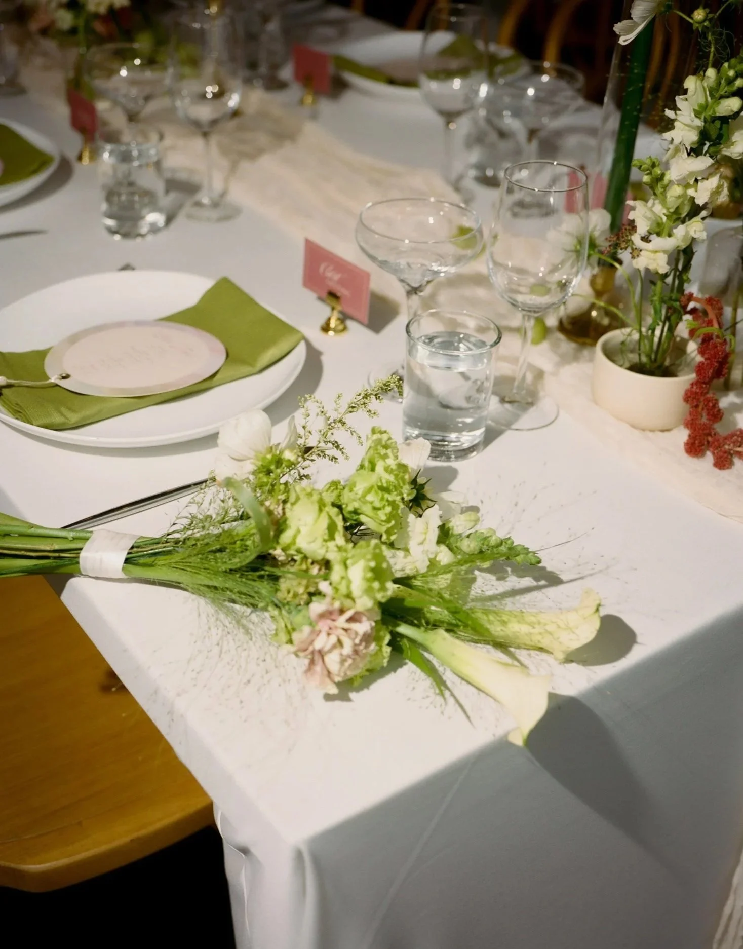 A formal dining table decorated with floral arrangements, glassware, and place settings. The table has a white tablecloth with a smaller green cloth and white plates, and is adorned with flowers and greenery.