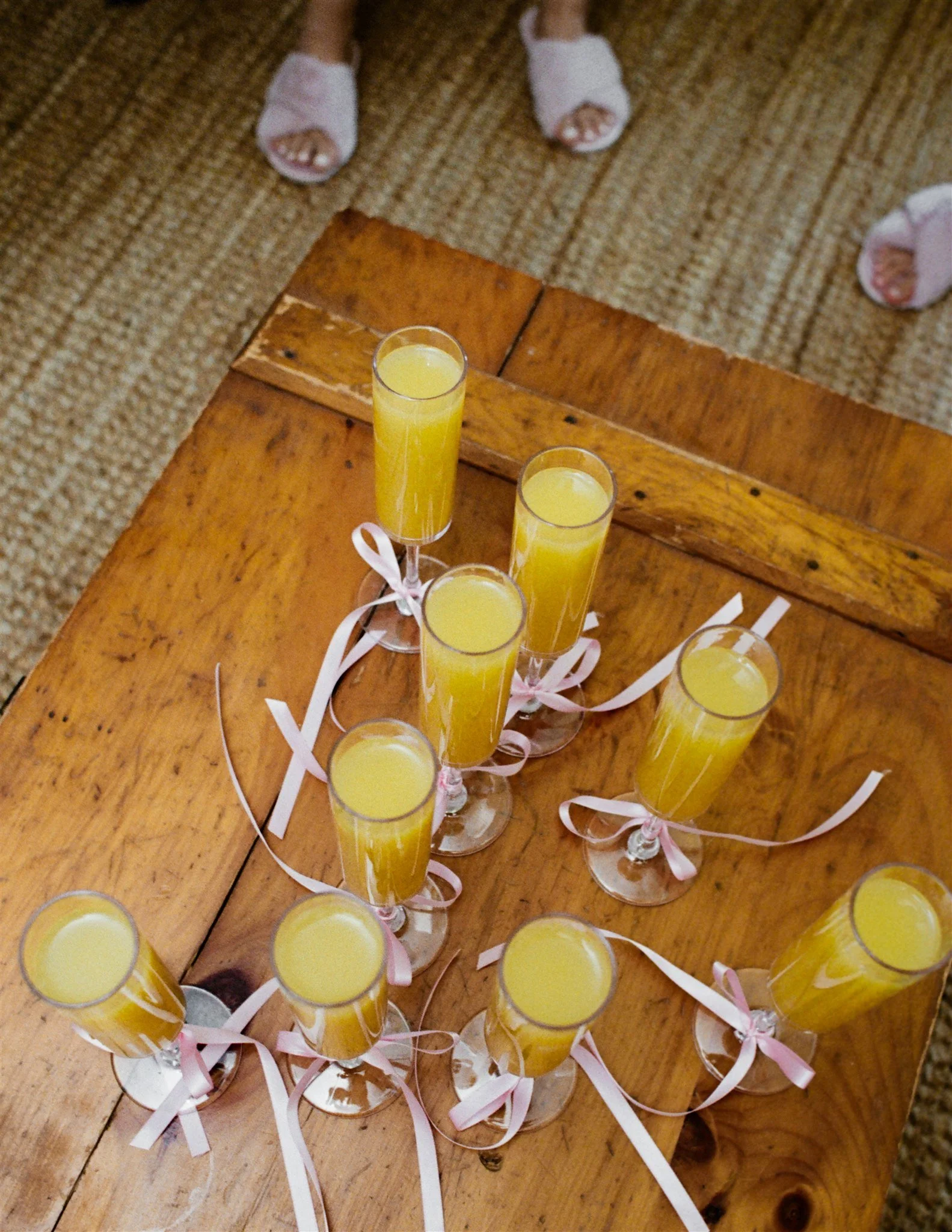 A group of glasses filled with yellow-orange juice, decorated with pink and white ribbons, placed on a wooden table. The background shows a carpeted floor and a person's feet in white slippers.