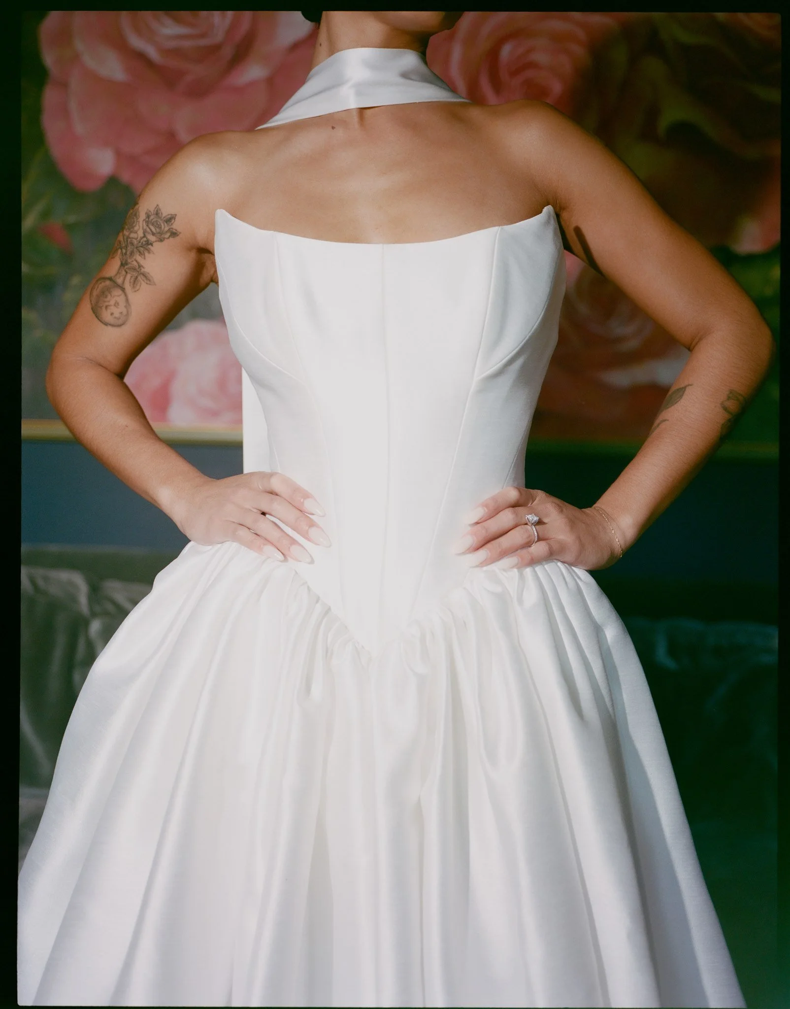 A woman in a white dress with a high collar, with tattoos on her arms, standing with her hands on her hips. The background features large pink roses.