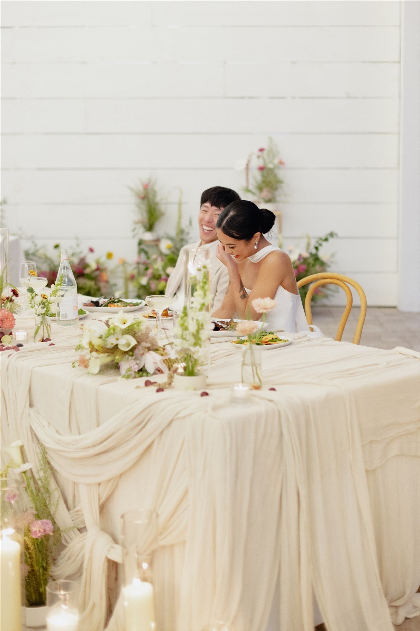 A wedding reception with a long table decorated with white flowers, candles, and draped fabric. Two people are sitting at the table, laughing and smiling, with a white wooden wall and some potted plants behind them.