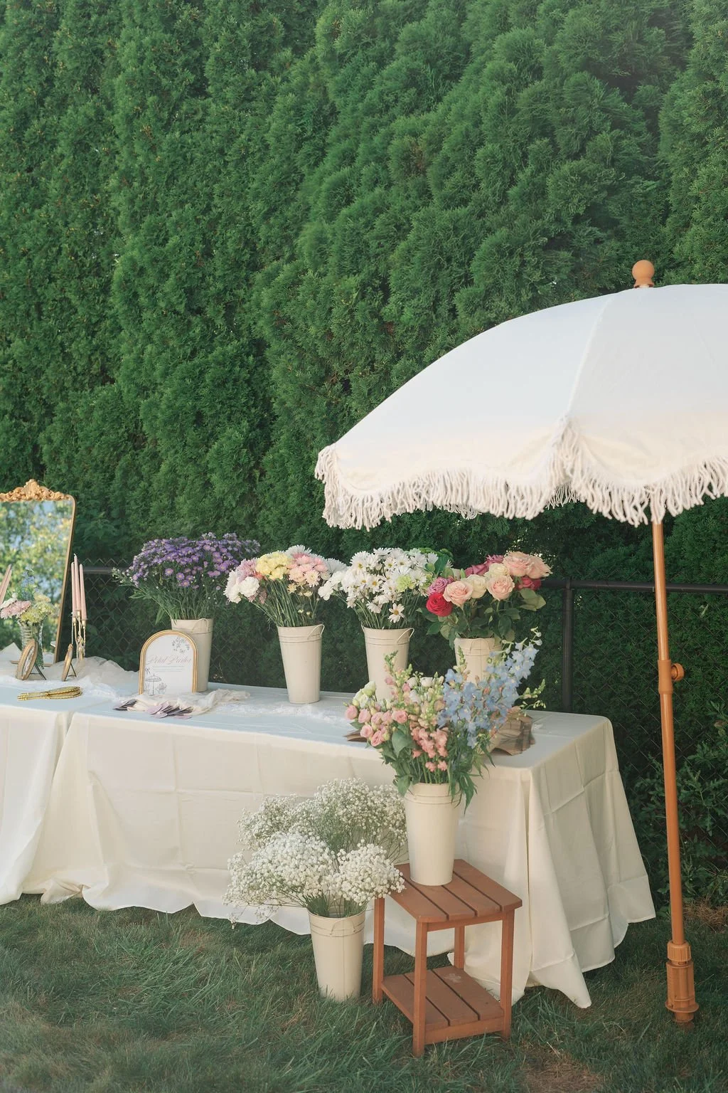 Outdoor table decorated with pastel flowers in white vases, a white tablecloth, and a large white parasol, set against a backdrop of green trees.