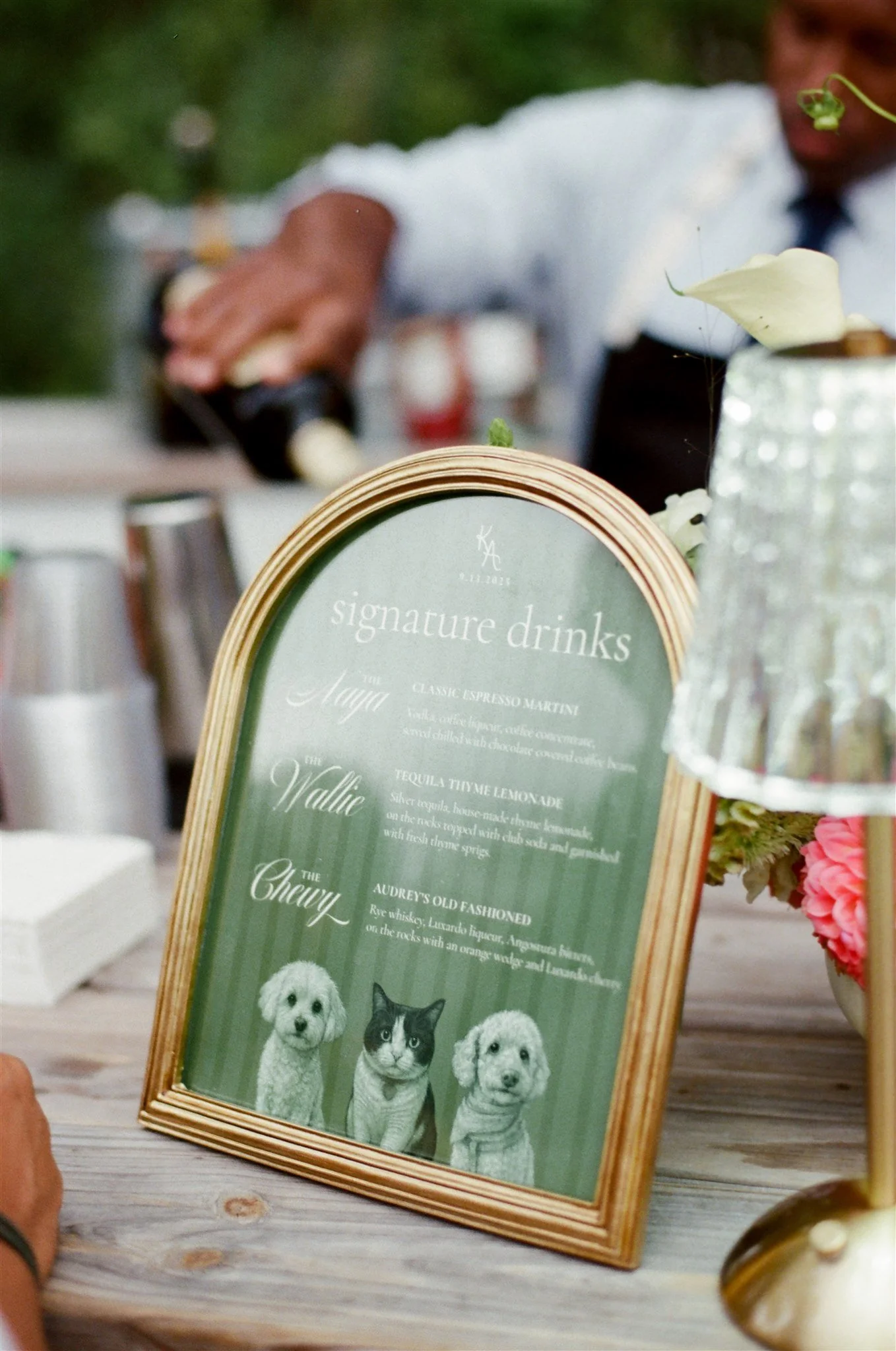 A framed sign displays drink options with illustrations of two dogs and a cat at a celebration or event. A person is pouring a drink in the background.