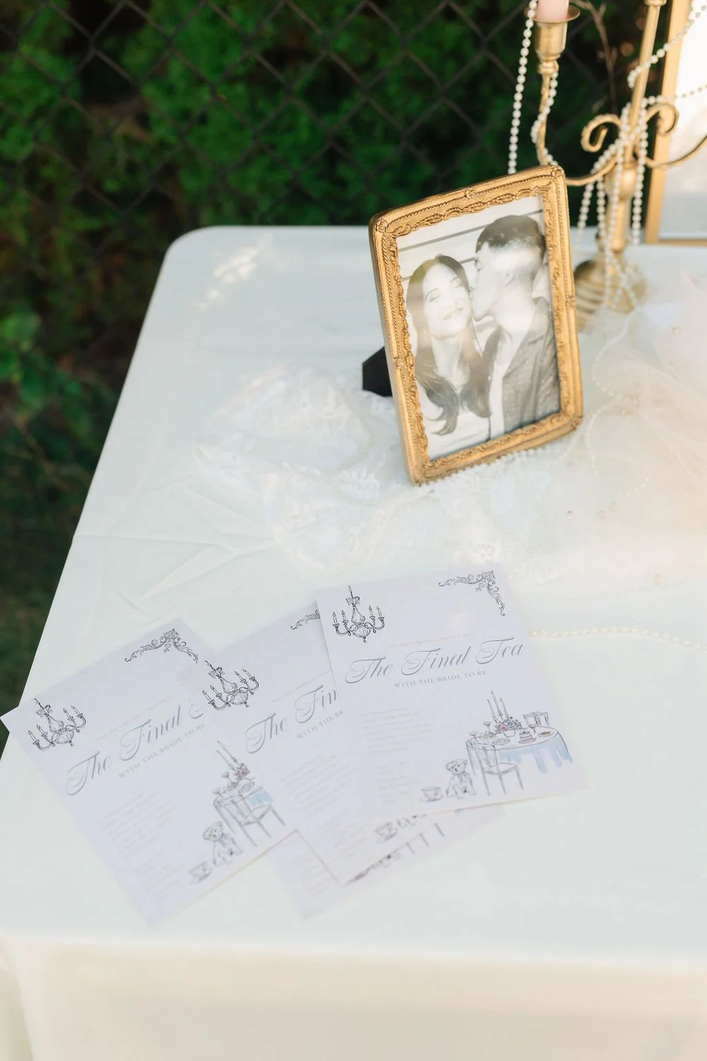 A framed black and white photograph of a smiling woman and man on a white table. The table also holds three printed programs titled "The Final Tea," with elegant decorative elements, placed overlapping each other. A candelabra with beads and candles 