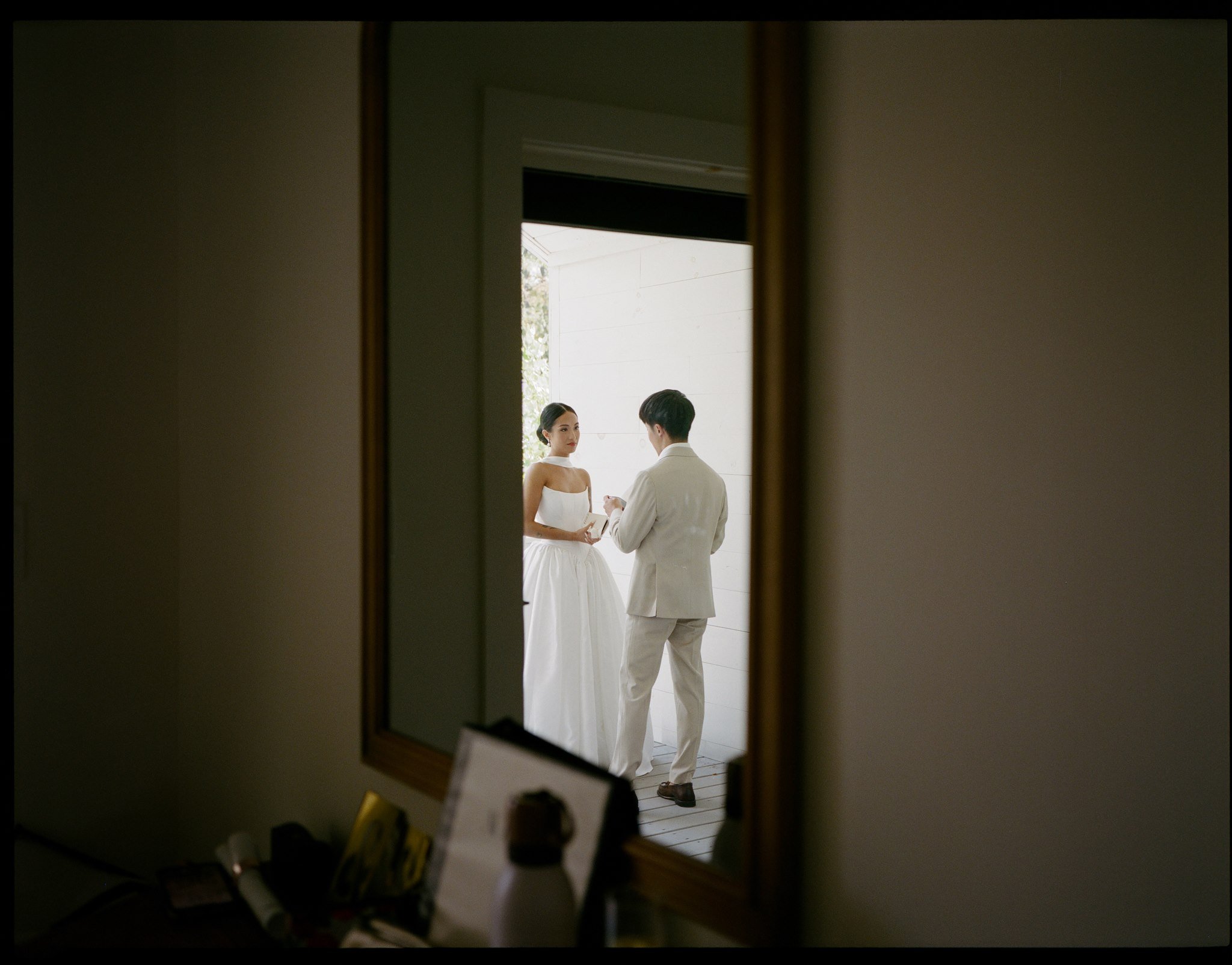 A reflection of a couple in wedding attire, standing outside near a white wall, seen through a mirror. The reflection shows the bride in a white dress and the groom in a white suit, with the bride looking at the groom.