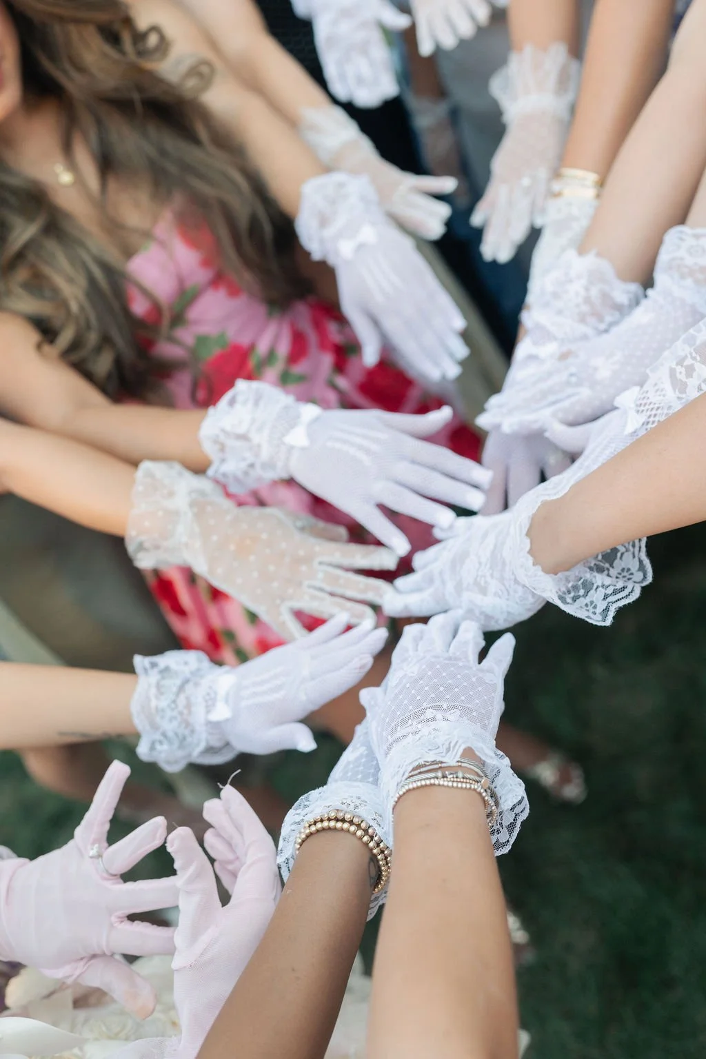 Multiple women wearing white lace gloves and accessories, with some women in floral dresses, gathered together with their hands extended towards the center.