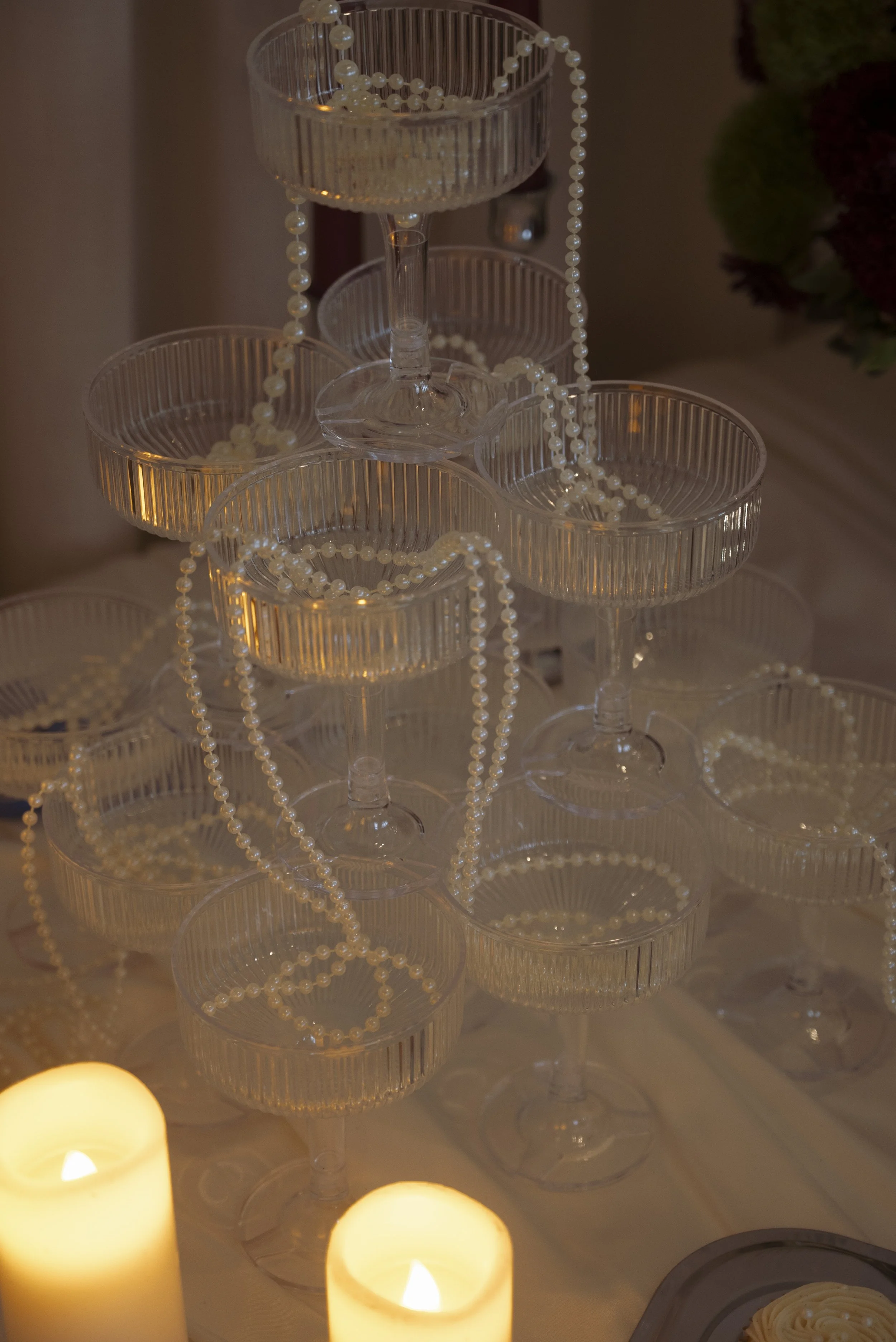 Empty glass dessert cups arranged on a stand with strands of pearl beads draped over them, and candles in the foreground illuminating the scene.
