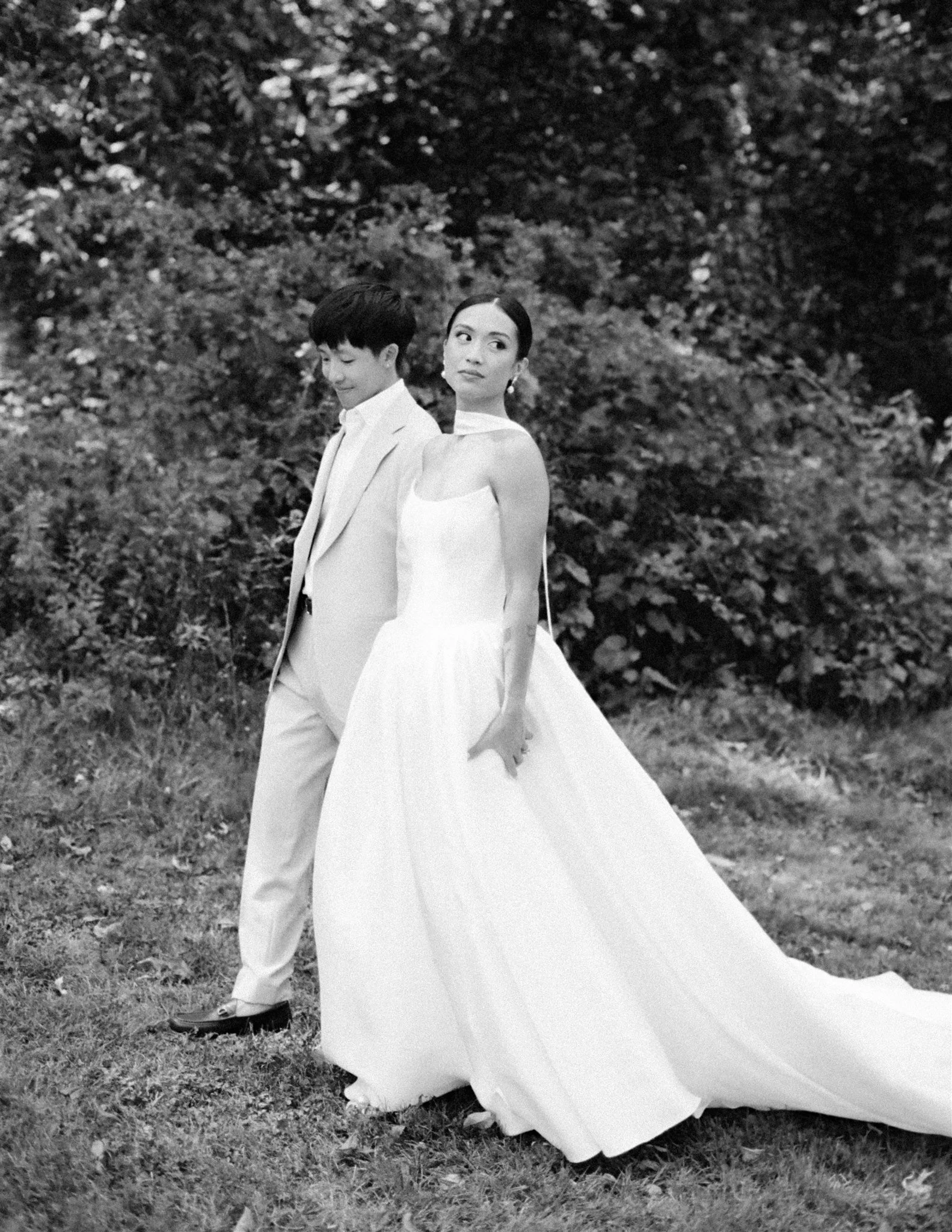 A black and white photograph of a man and woman standing outdoors in a natural setting with trees and grass. The woman is wearing a white dress and looking over her shoulder, while the man is dressed in a light suit and looking downward.