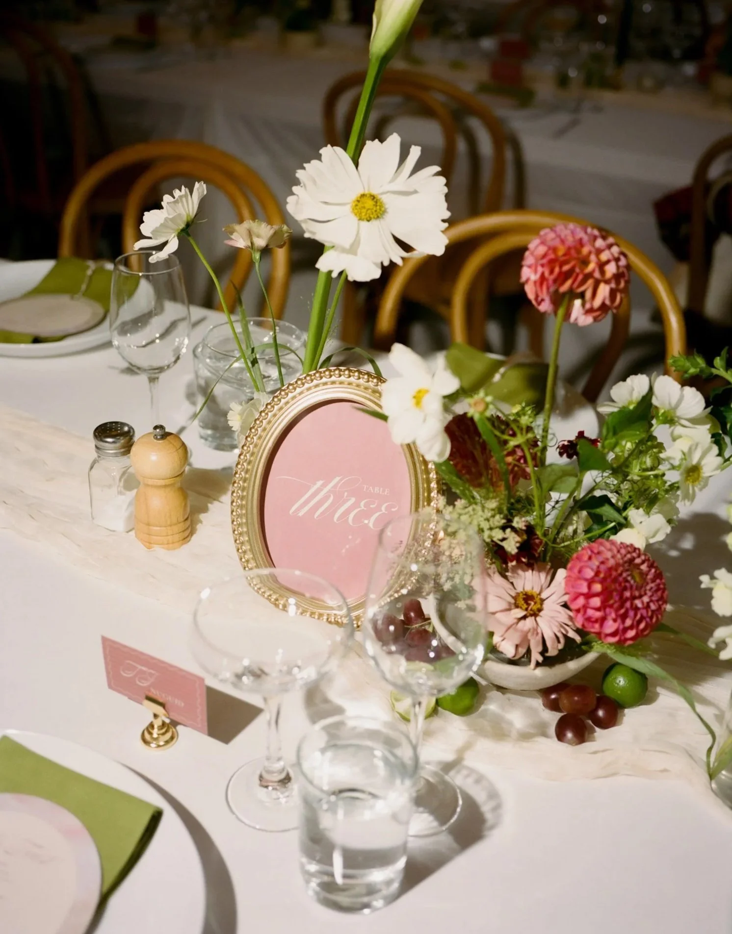 Table with floral centerpiece, pink and white flowers, wine glasses, salt and pepper shakers, and a pink table sign at a formal event or wedding reception.