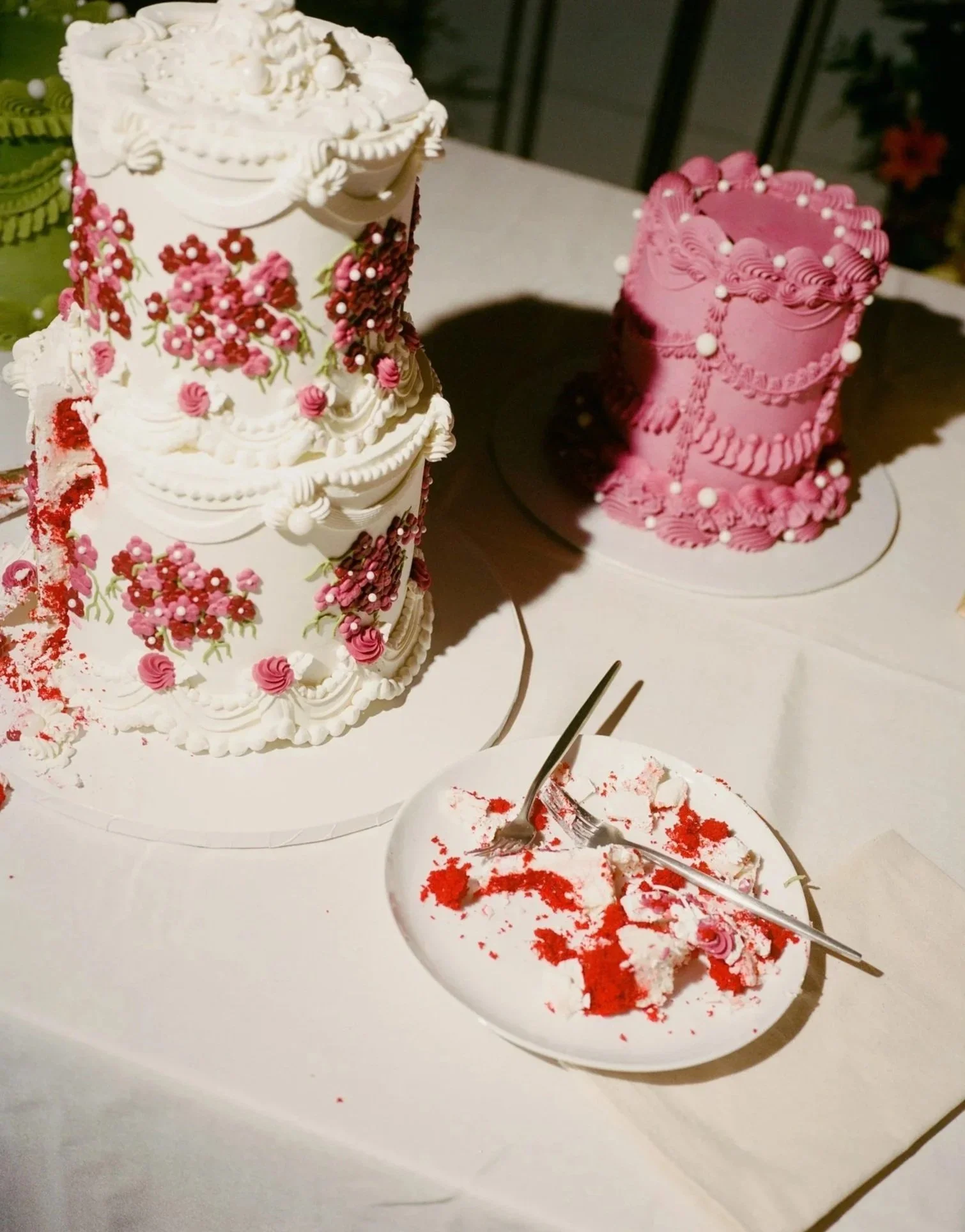 A white and pink decorated cake with floral and scroll designs, partially eaten on a white table, with a piece of cake in a white dish topped with red and white sprinkles and red crumbs, a silver fork, and a silver cake server.