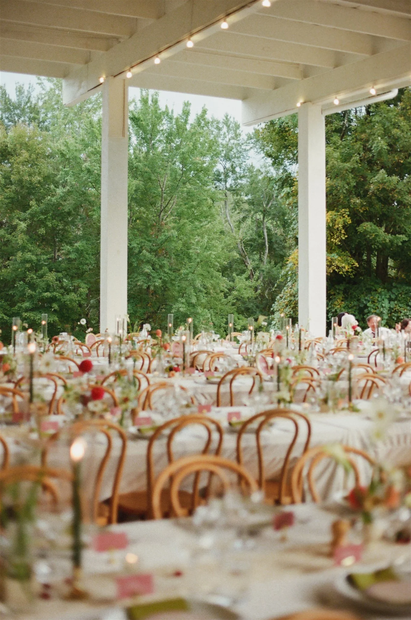 Outdoor reception area with long tables set with floral centerpieces and tall candles, under a porch with string lights and green trees in the background.