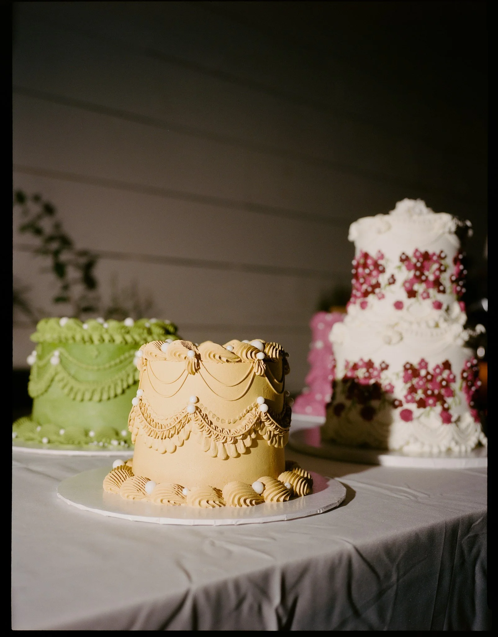 A three-tiered cake decorated with pink and white floral patterns and scalloped icing, placed on a white table with a gray tablecloth, with two other cakes in the background.