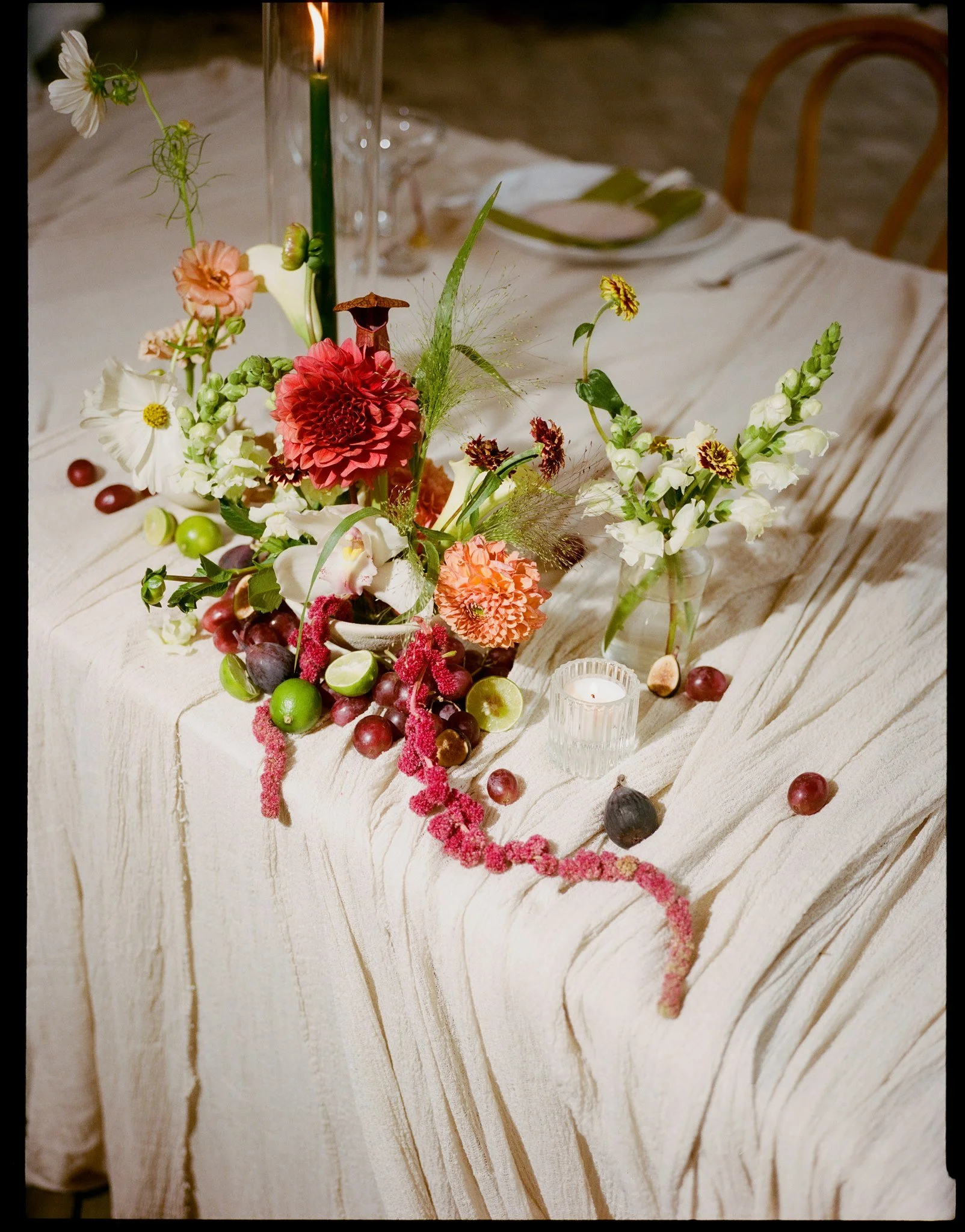 A table decorated with a floral centerpiece, including roses, lilies, and other flowers, with scattered grapes, a candle, and a white tablecloth.