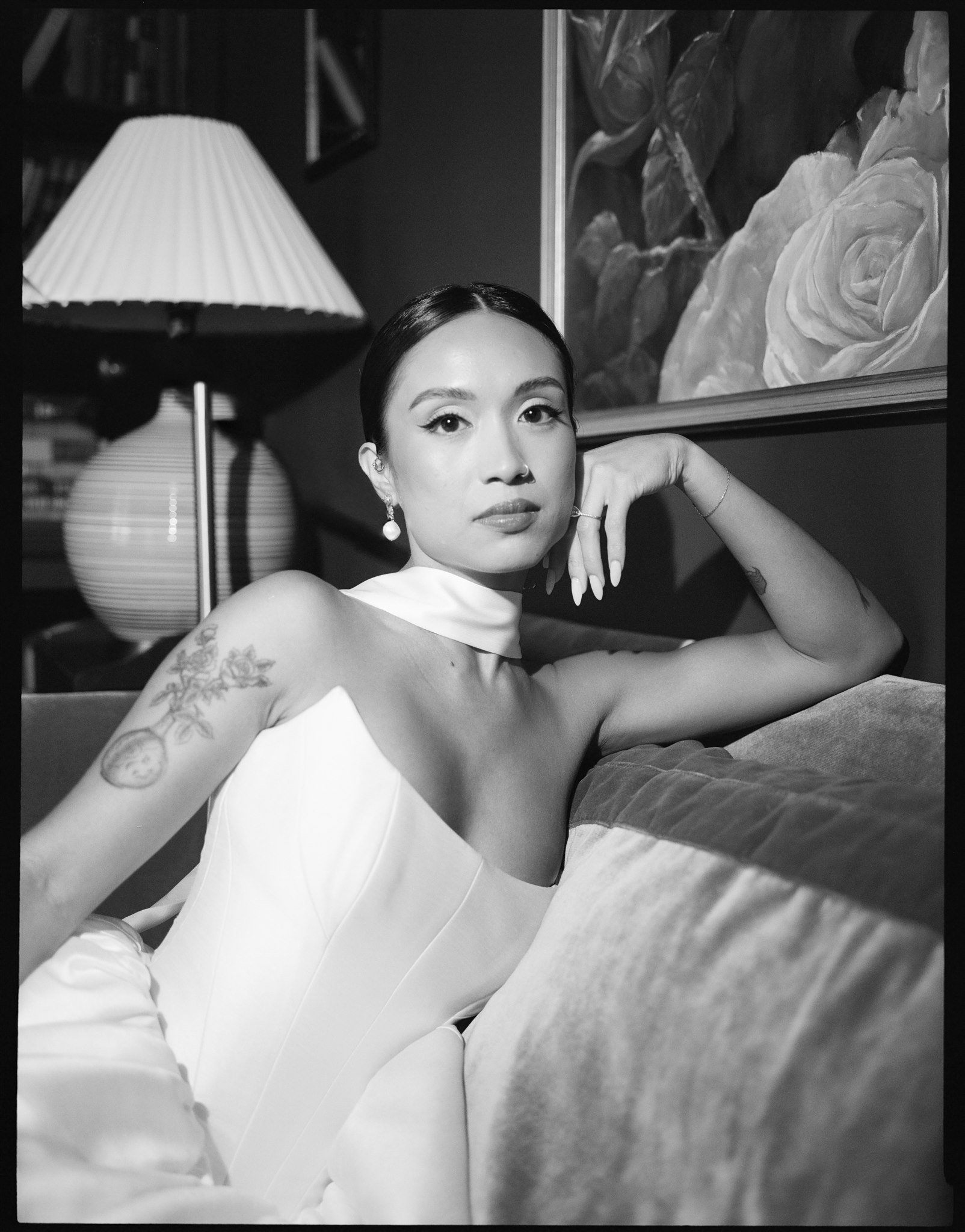 Black and white photo of a woman with dark hair slicked back, wearing earrings, and a dress with a sleeve covering her left shoulder, resting her arm on a surface with a textured fabric, in front of a painting with roses and a lamp in the background.