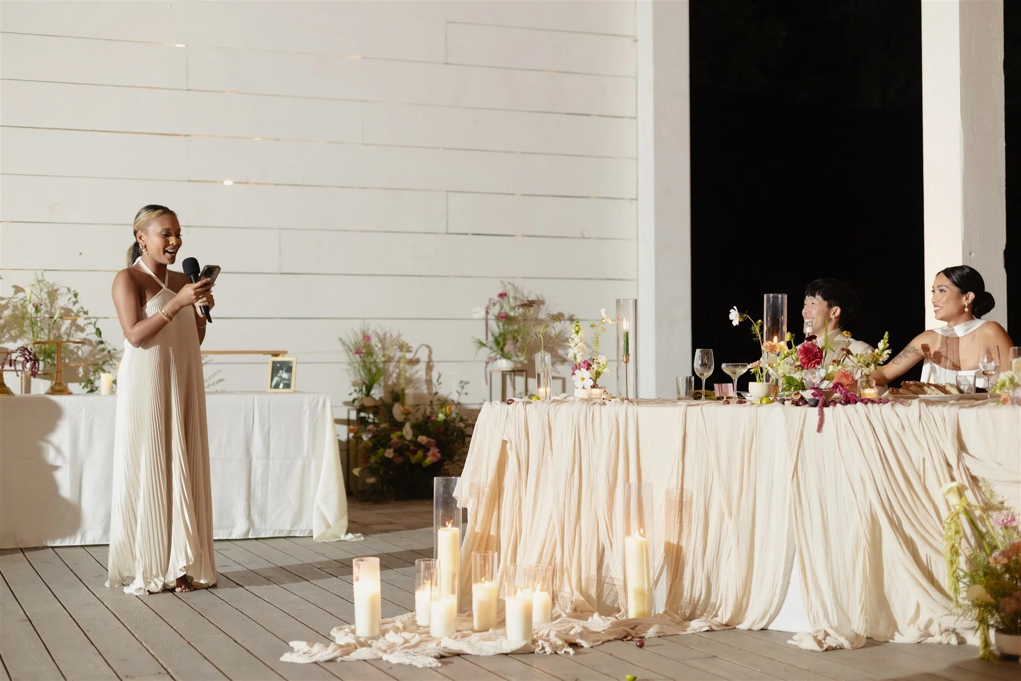 Woman in a beige dress giving a speech at a wedding reception with floral arrangements and candles on the table.