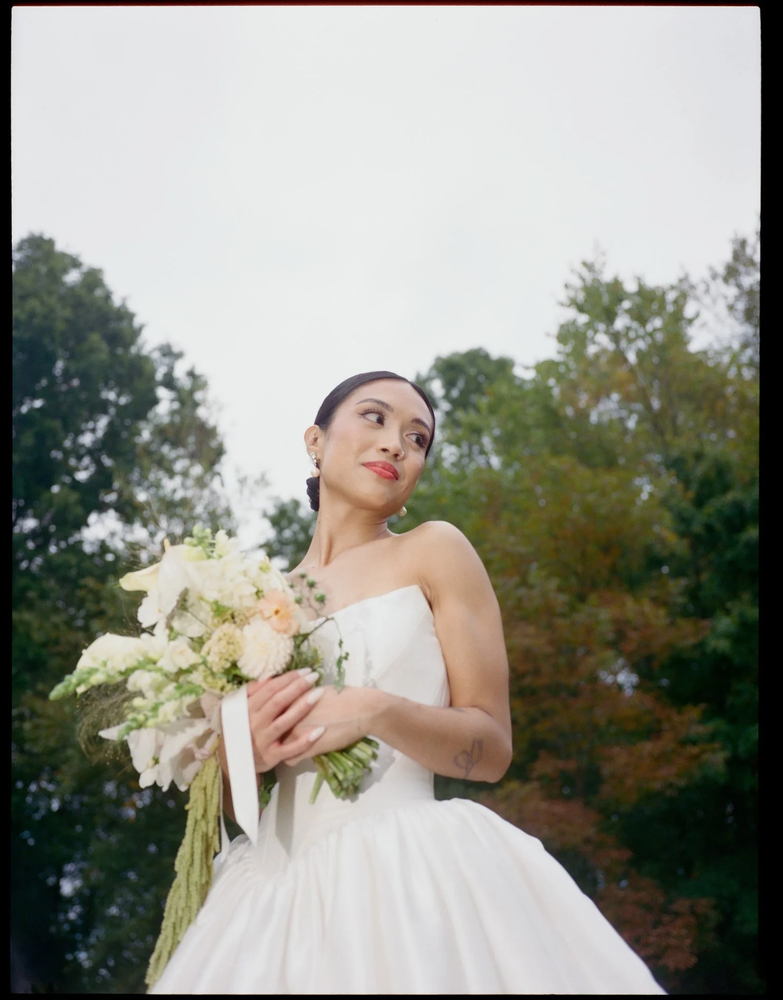 A woman in a white wedding dress holding a bouquet of flowers outdoors with trees in the background.