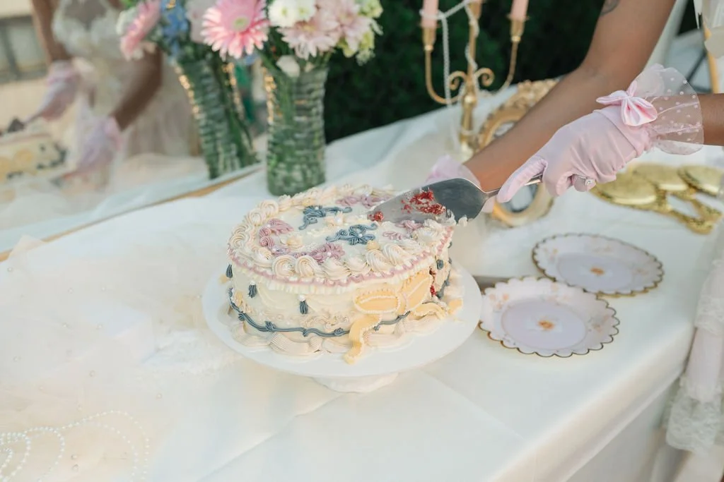 A person wearing pink gloves is cutting a decorated birthday cake with floral and ribbon decorations. There are vintage china plates, a white tablecloth, and a floral centerpiece in the background.